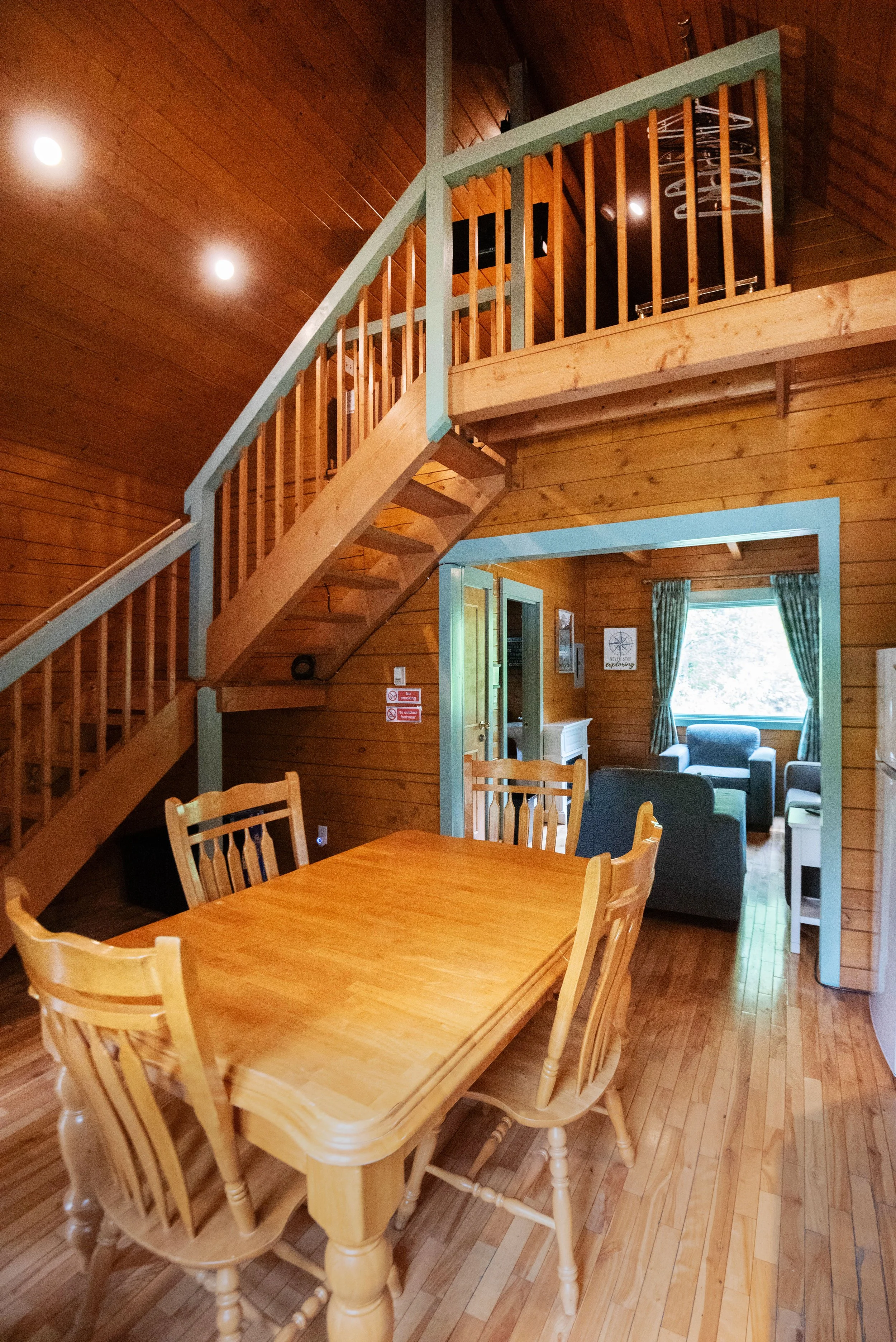 Interior view of a wooden cabin with a dining table and chairs in the foreground, a second seating area with sofas in the background, and a staircase leading to an upper level. Wood paneling covers the walls and ceiling, with framed pictures and sign
