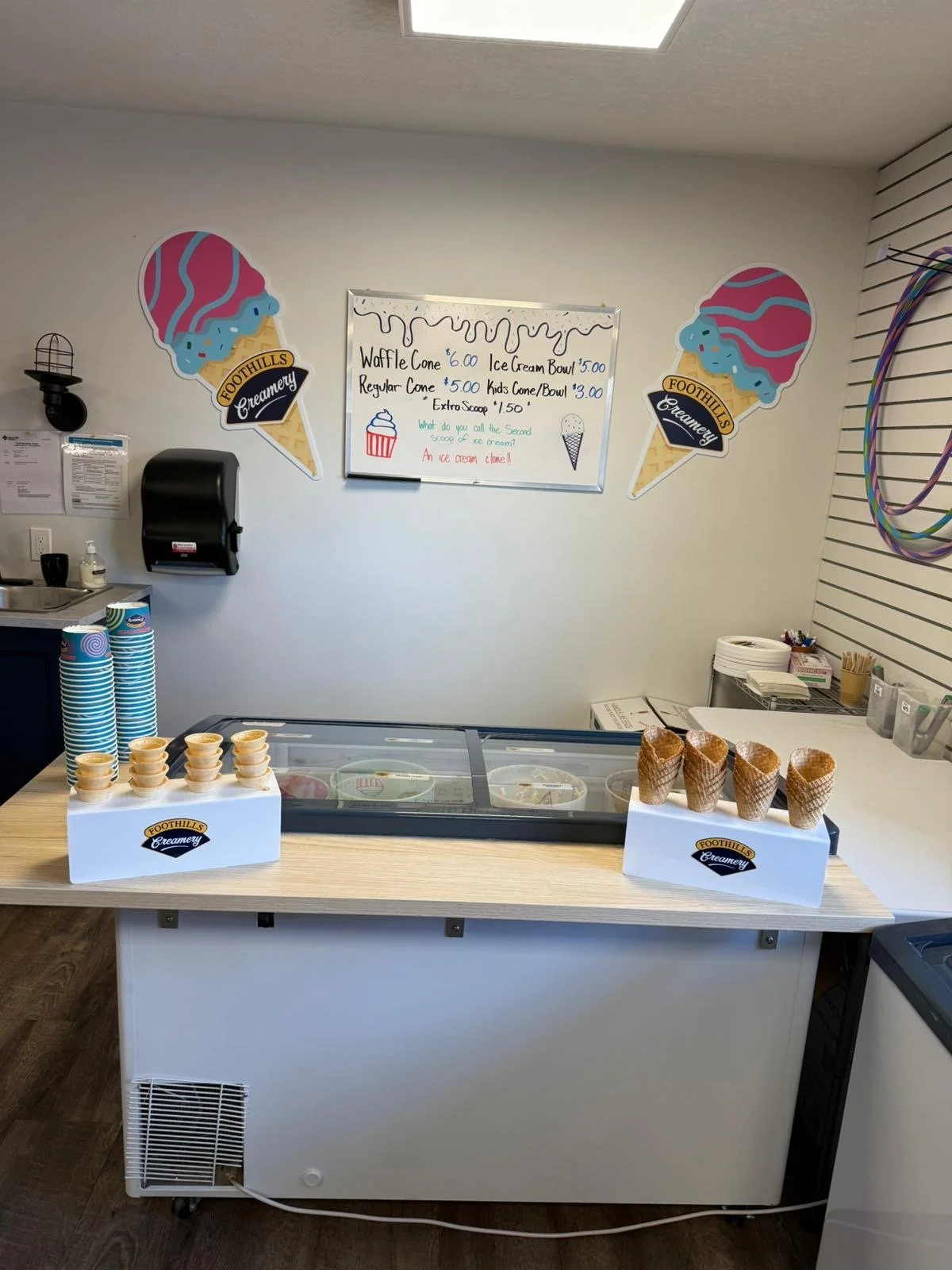 Ice cream shop counter with two display boxes of ice cream cones, a whiteboard menu, and wall decorations including ice cream cone cutouts and hula hoops.