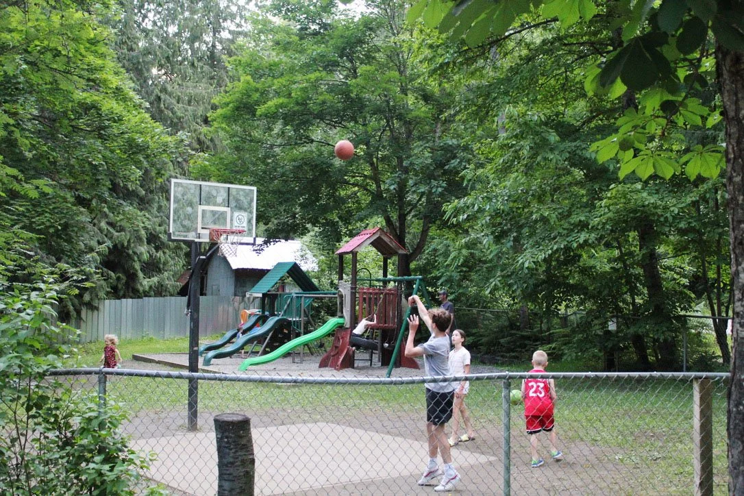 Children playing basketball on a court in a park with trees and a playground in the background.