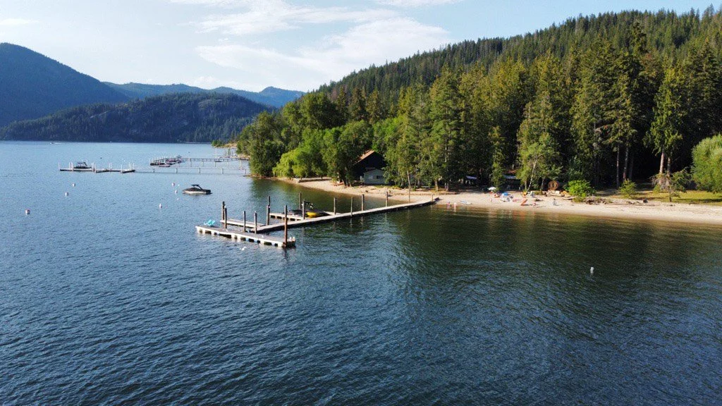 Scenic view of a lake with a small sandy beach and a boat dock, surrounded by dense evergreen trees and mountains in the background.