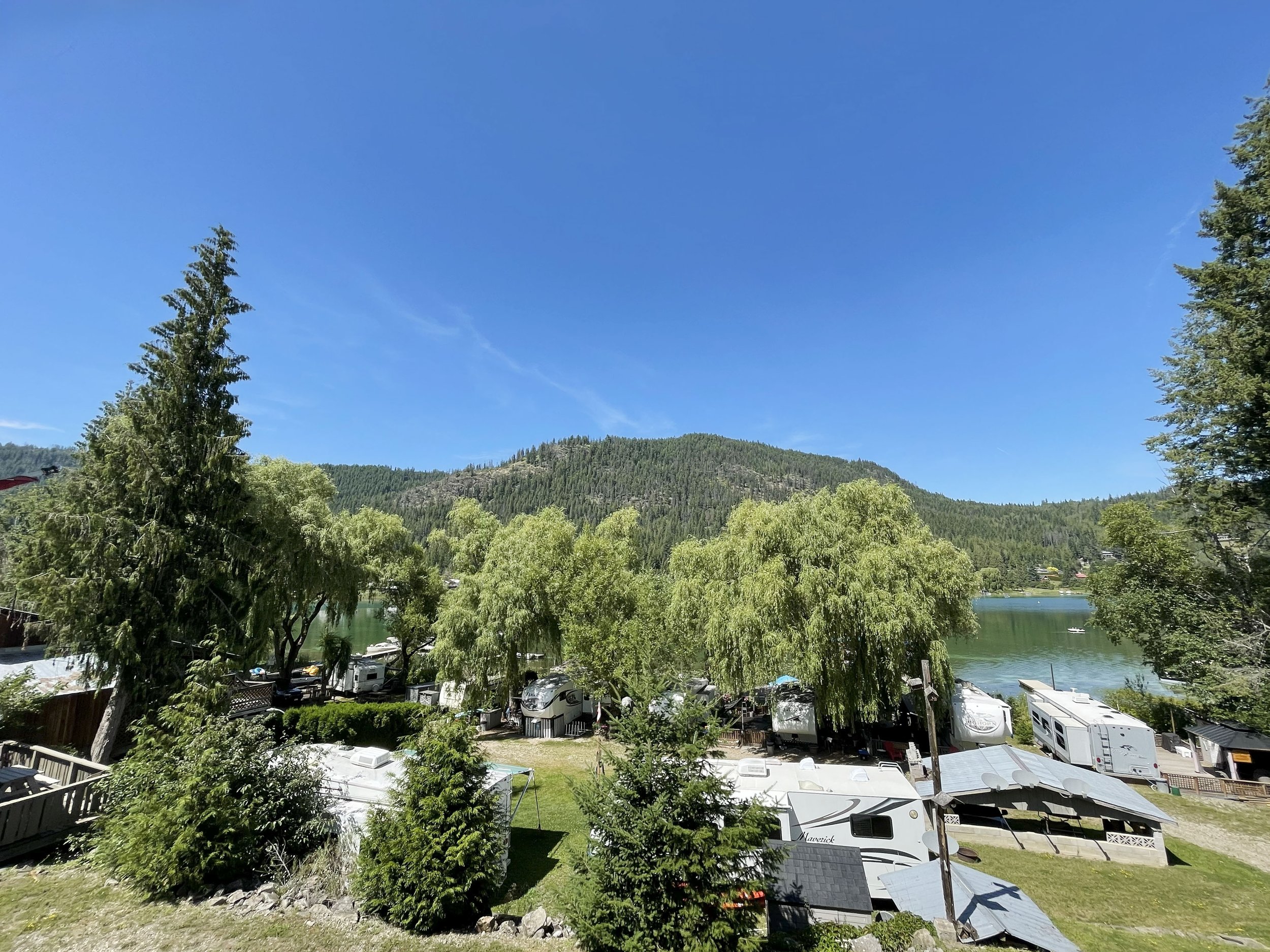 A scenic landscape featuring a lake, lush green trees, a forested hillside, and a clear blue sky. There are RVs and boats near the shoreline, indicating a recreational area.