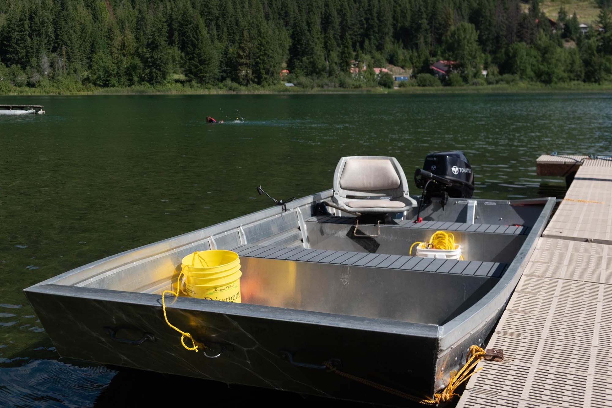 A lakeside dock with a small aluminum boat tied to it, containing yellow buckets and a rope, with a boat engine at the rear, against a backdrop of trees and mountain houses.