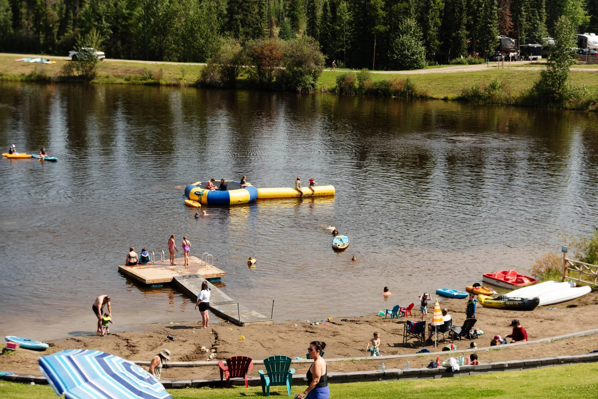 People enjoying a sunny day at a lake with boats, kayaks, and a floating water trampoline, surrounded by green trees and a grassy area with a parking lot in the background.