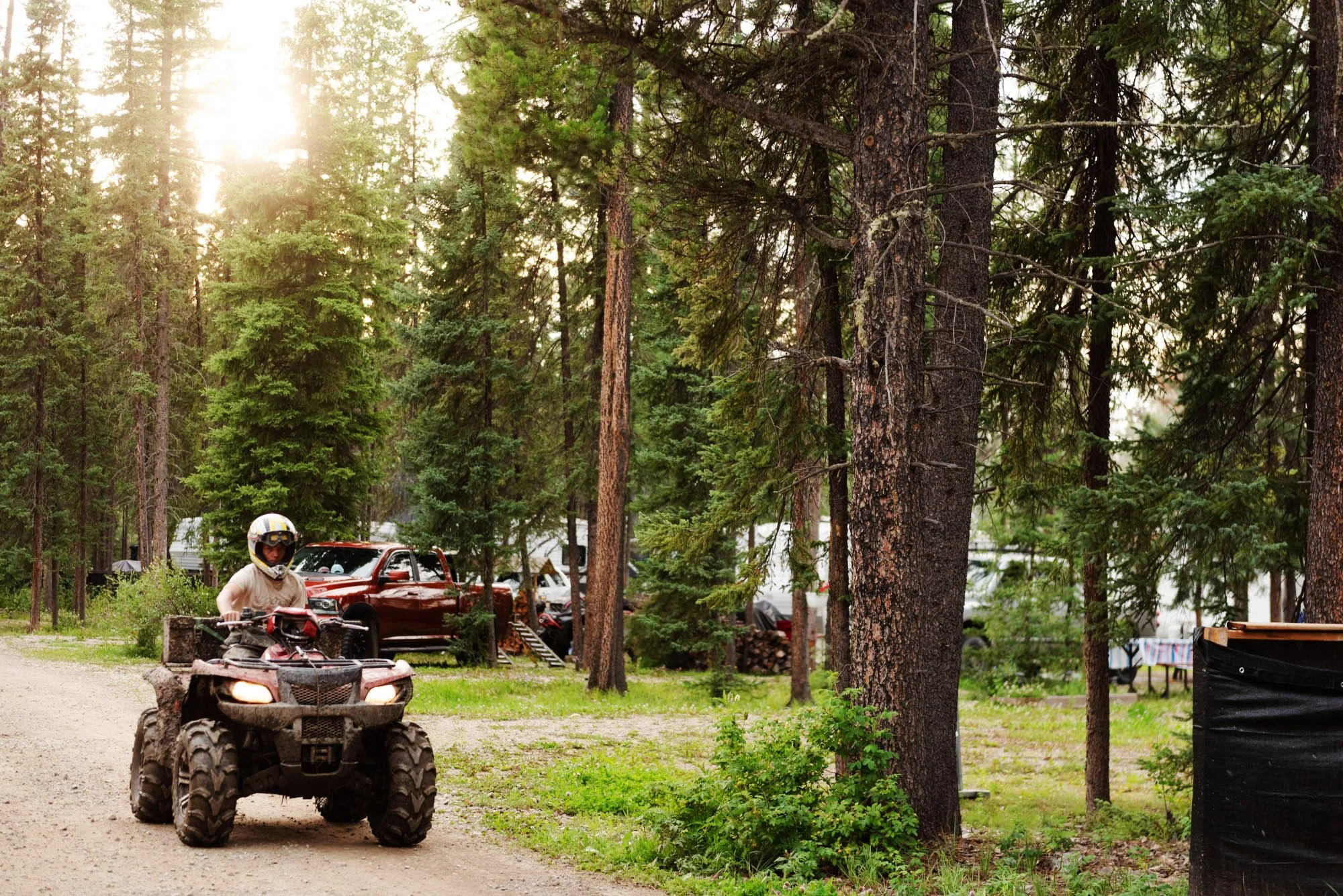 A person riding a four-wheeler ATV on a dirt path in a wooded area with tall trees, sunlight filtering through the branches, and parked cars and a red pickup truck in the background.