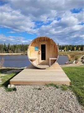 A round wooden structure resembling a barrel sauna or tiny house on a dock by a lake, with a screened door and small window, under a cloudy sky.