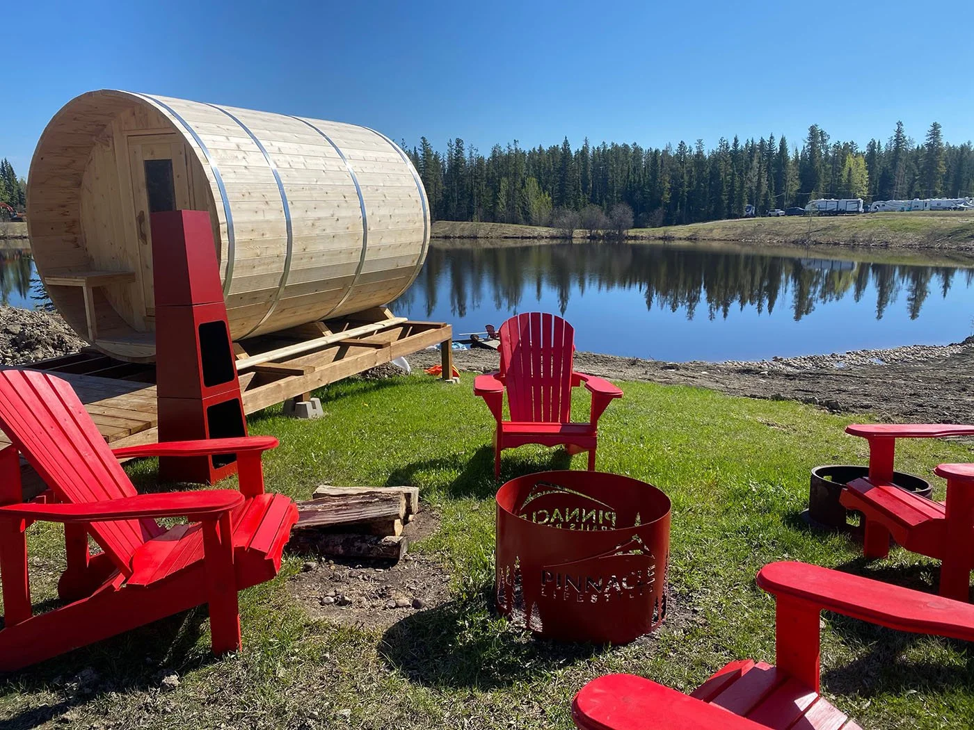 Red Adirondack chairs and fire pit arranged on grass near a small pond, with a wooden barrel-shaped sauna and forested trees in the background under a clear blue sky.