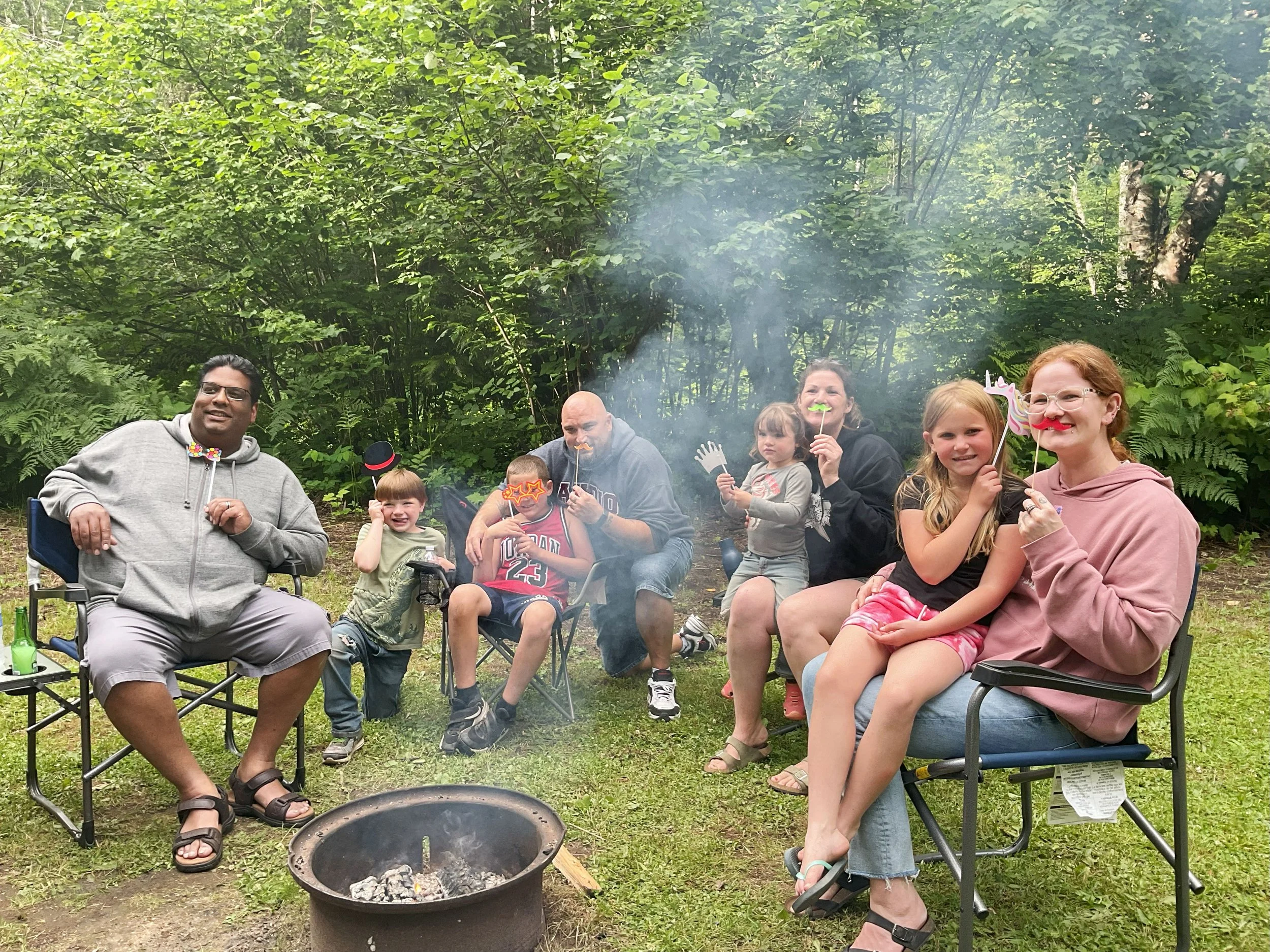 Family and friends gathered outdoors in a forest clearing, sitting around a campfire with light smoke rising. Some people hold festive photo props and wear casual clothing, including a woman with glasses and red hair, children, and adults of diverse backgrounds, enjoying a relaxed social gathering.