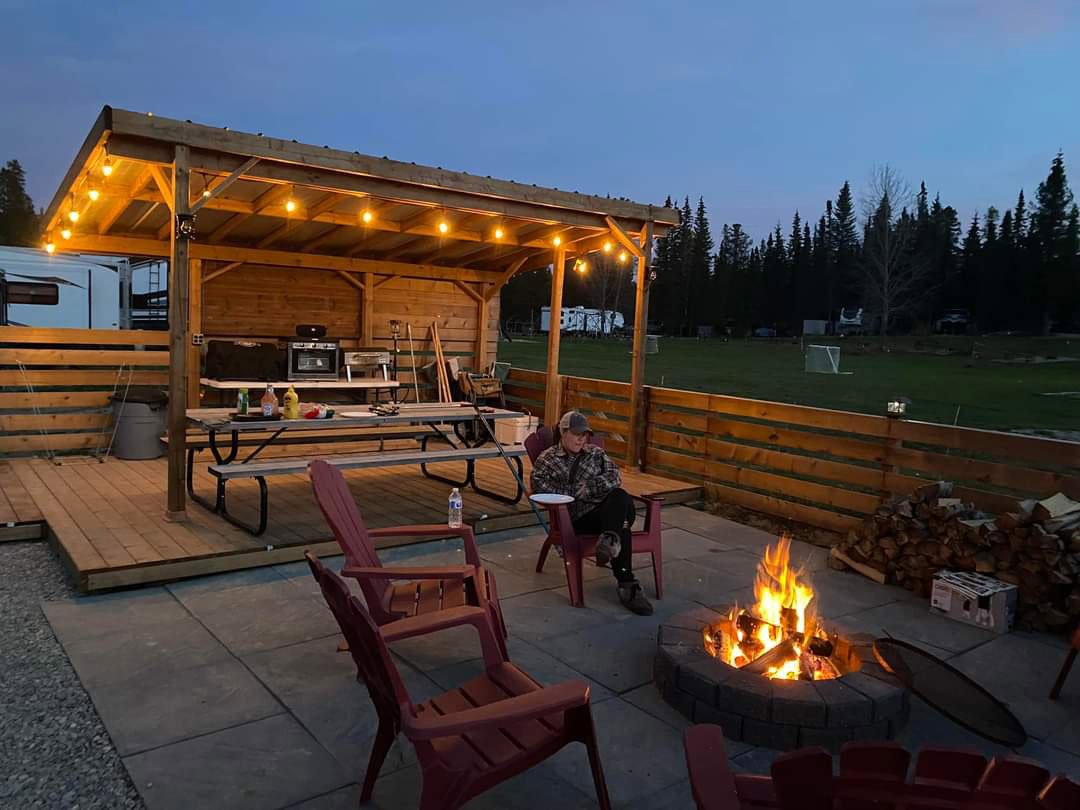 Outdoor patio with a fire pit, red chairs, and a wooden shelter with string lights. A person is sitting in a red chair near the fire, and the shelter has a picnic table with bottles and food on it, and a grill in the background.