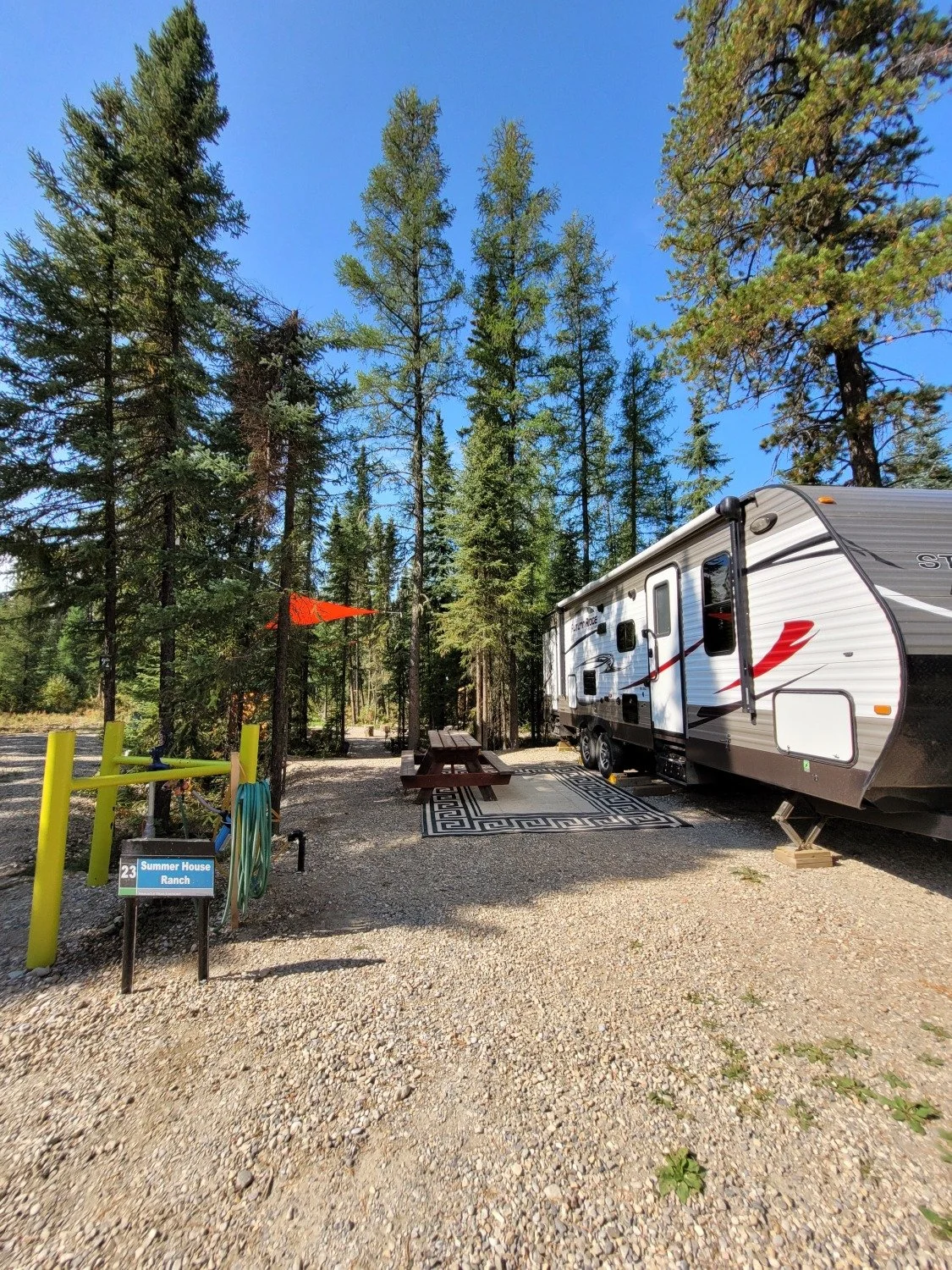 A campsite with a camper trailer parked on gravel, a picnic table with a rug, surrounded by tall pine trees, under a blue sky. There's a small sign that reads 'Summer House Ranch' and a hose hanging on a yellow post.