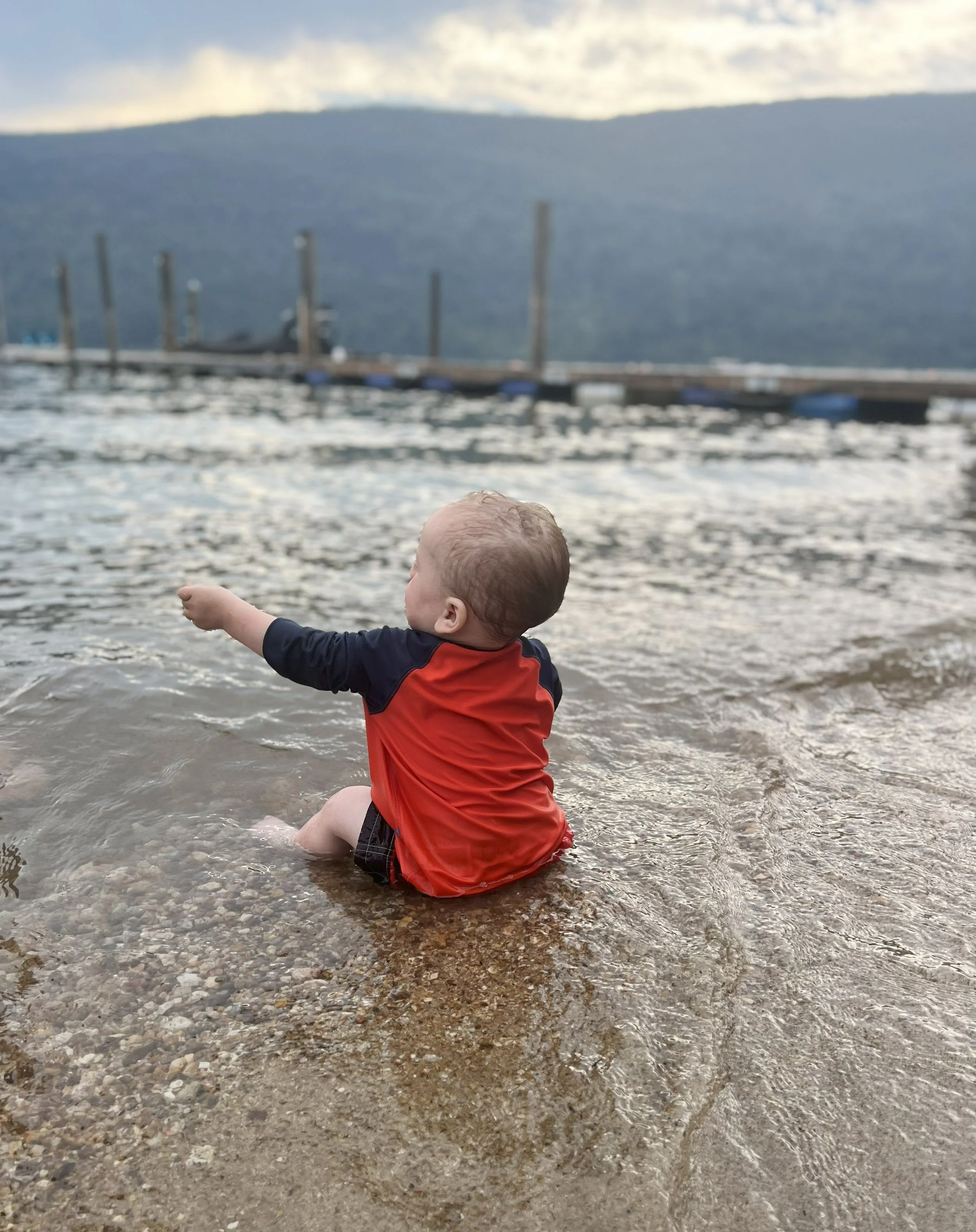 A young child sitting in shallow water at the edge of a lake or ocean, wearing a red and black long-sleeve shirt with short hair. In the background, there is a distant dock or pier with posts and a boat, and a mountains and cloudy sky.