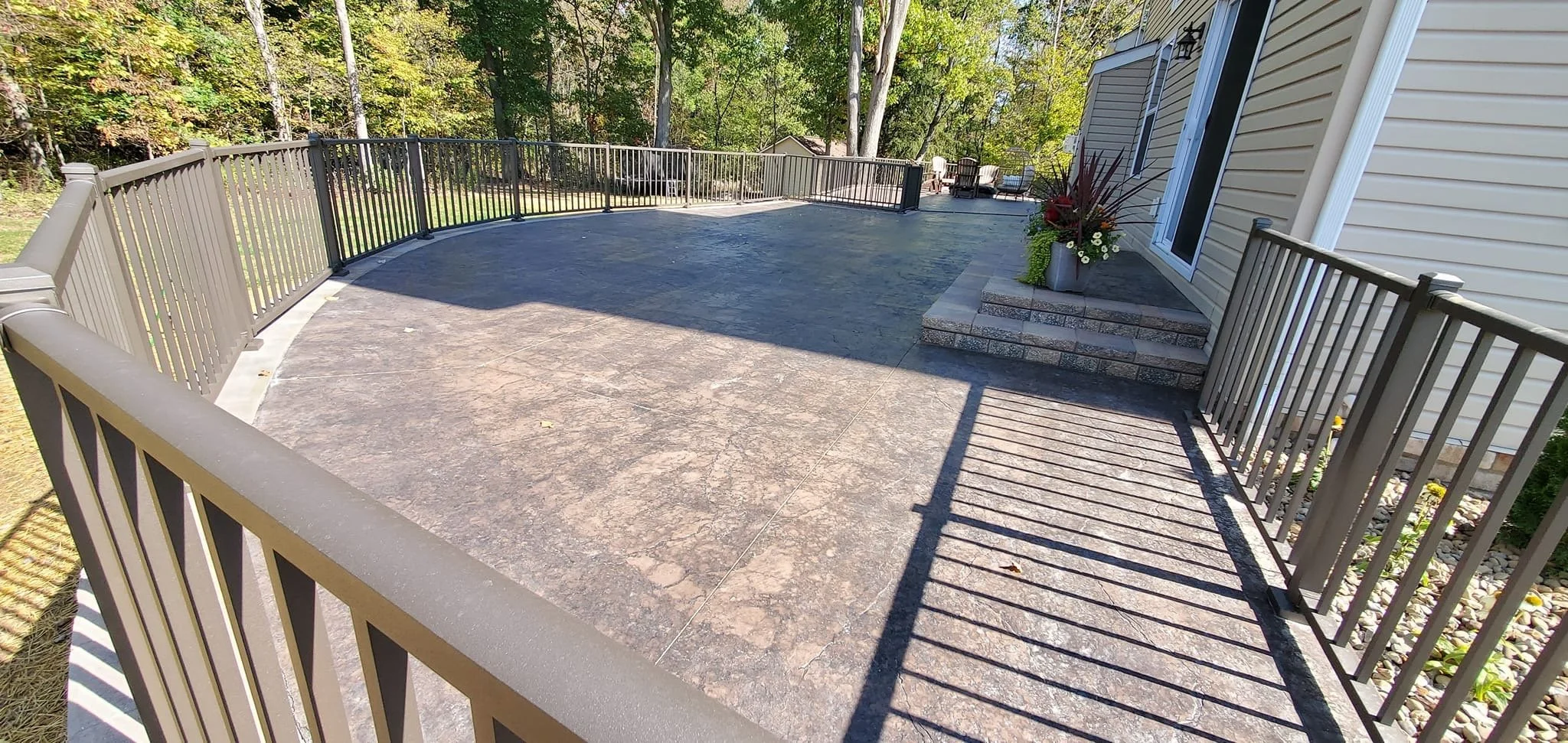 View of a spacious outdoor patio with a textured concrete surface, surrounded by a metal railing. There are steps leading up to the house with flowers in planters, and a seating area further back near trees and foliage.