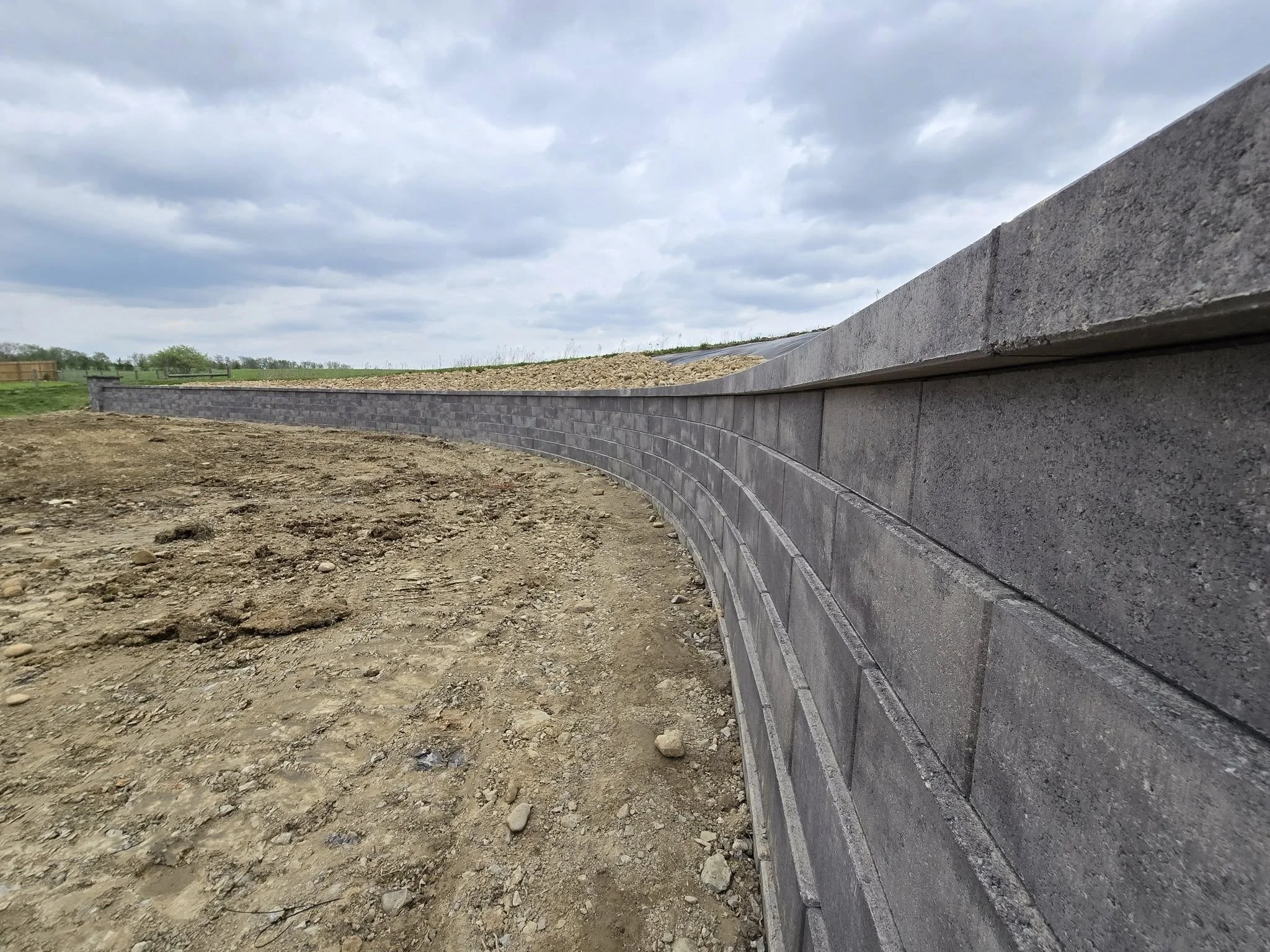 A curved concrete block wall in an outdoor construction site under cloudy sky, with dirt ground in the foreground and open land in the background.