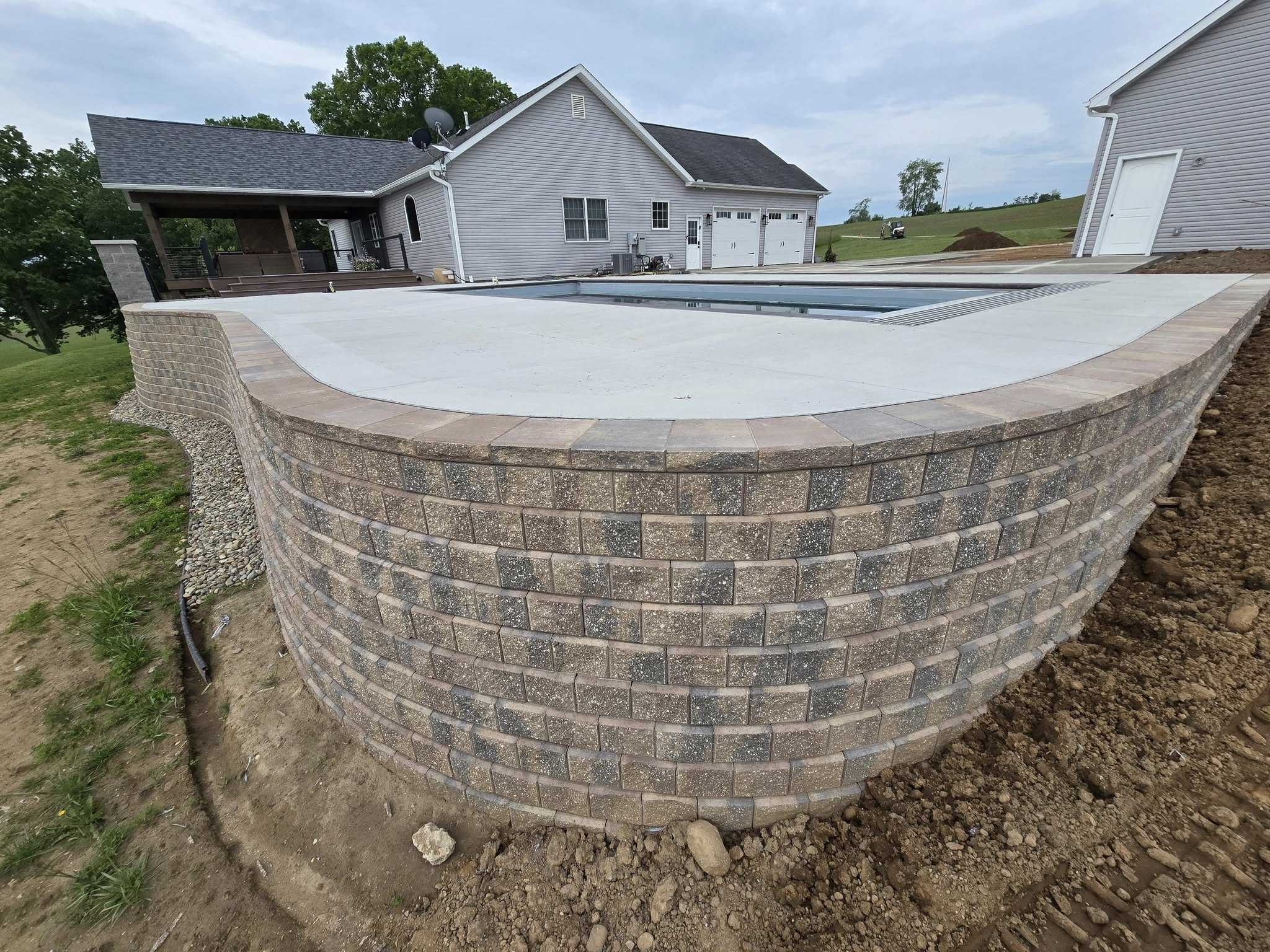 View of a backyard pool with a curved brick retaining wall and a paved deck, adjacent to a house with a covered patio, on a cloudy day.