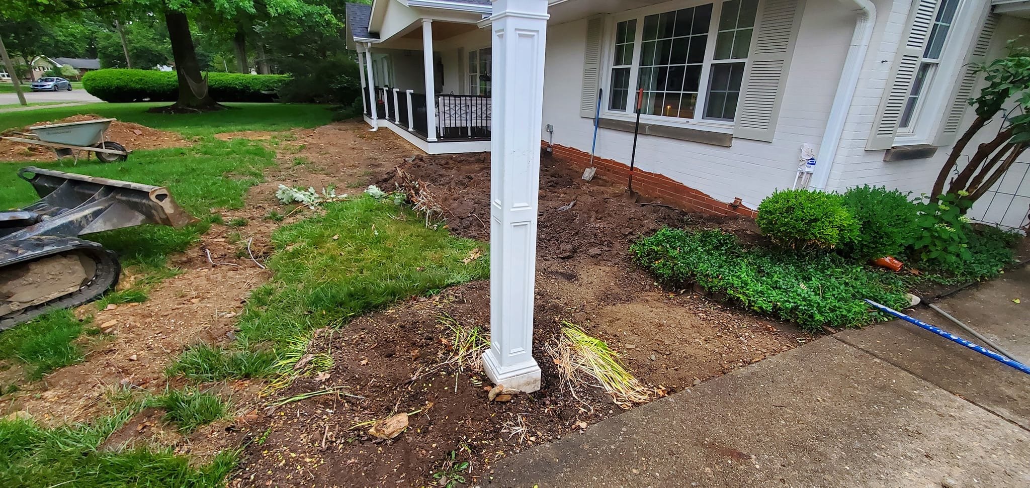 Front yard landscaping in progress with freshly turned soil, construction tools, and partially removed plants near a white house with an attached porch.