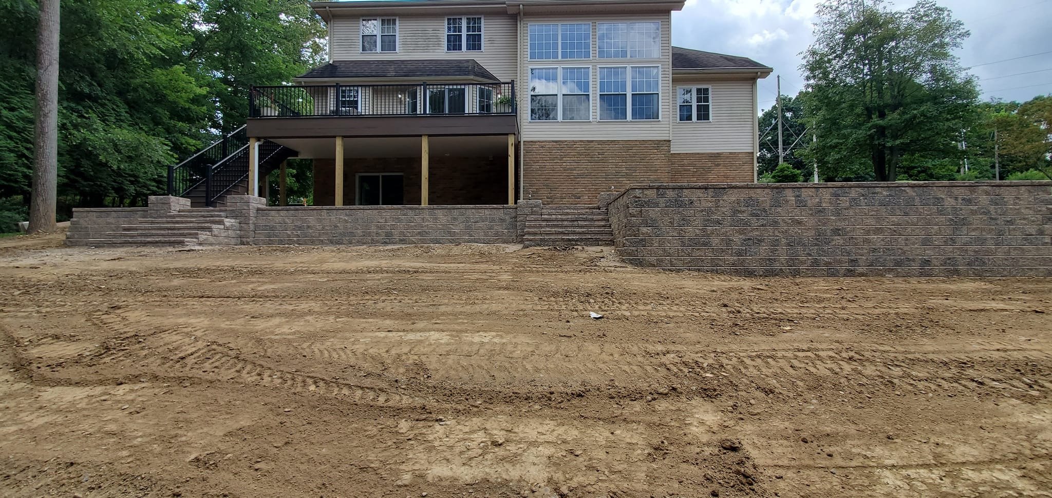 Backyard with dirt ground, staircases, and a retaining wall, attached to a multi-story house with large windows and a balcony.