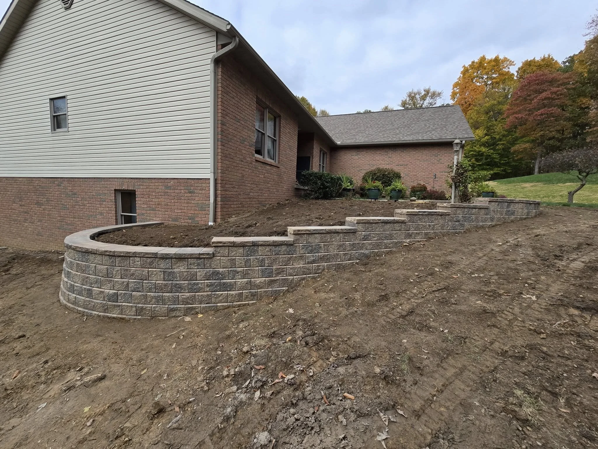 Backyard landscaping with a tiered retaining wall made of gray bricks, soil in front, and a house with a white and brick exterior in the background. There are potted plants near the house and a grassy area with trees showing fall colors.