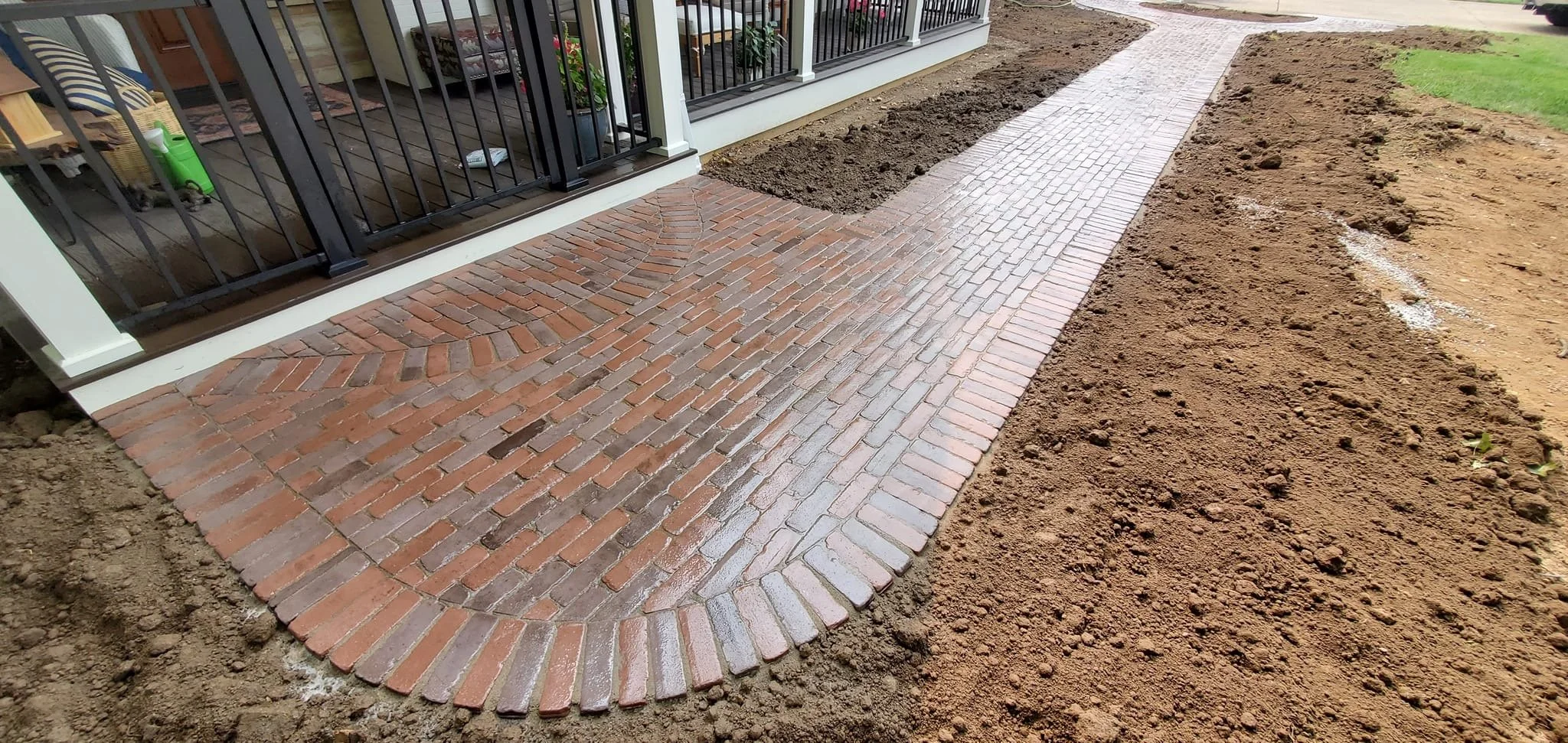 A newly paved brick sidewalk adjacent to a house with a porch, with dirt on either side waiting for landscaping.