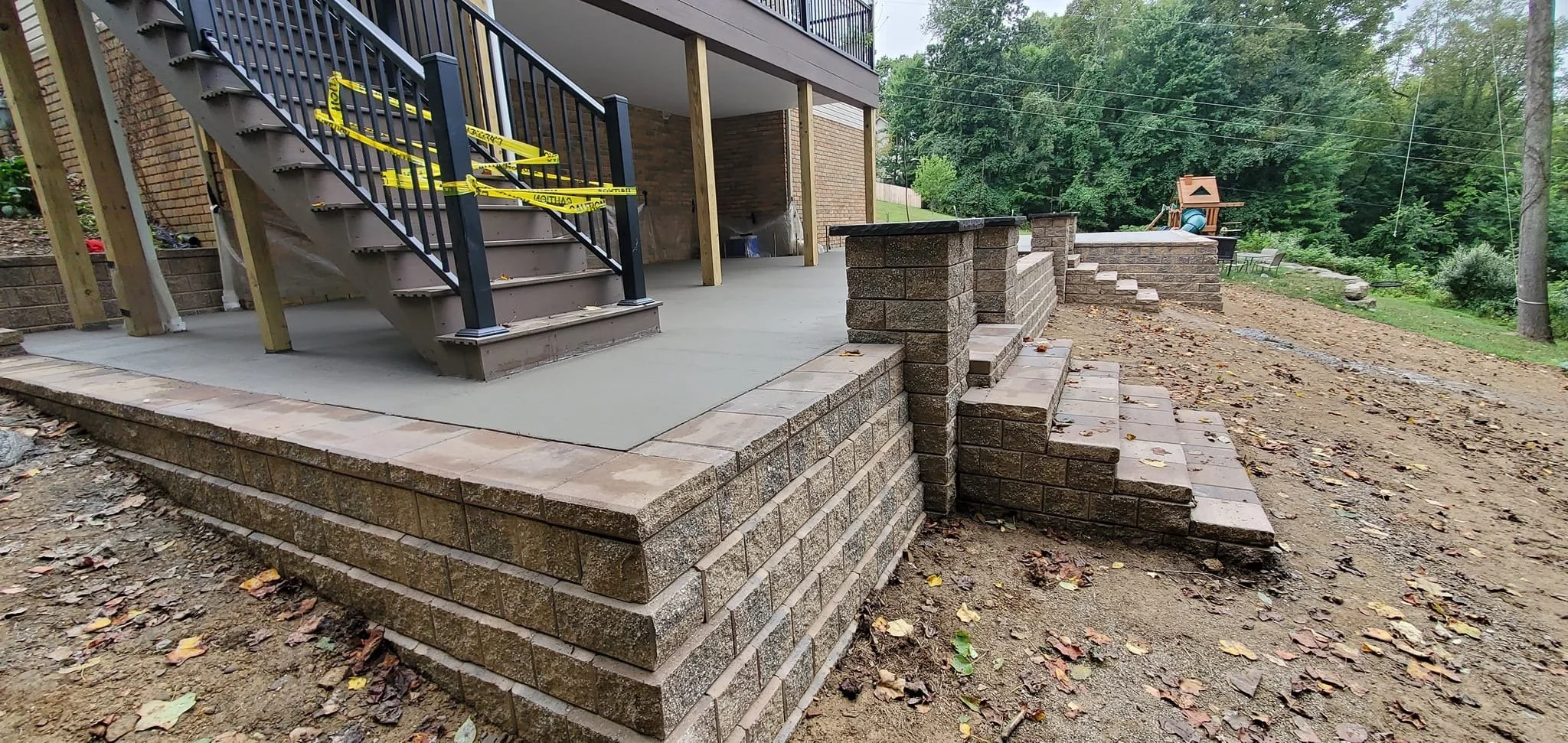 A newly constructed raised patio with brick steps and retaining walls in a backyard, with outdoor stairs leading to a deck, surrounded by trees and a dirt yard.
