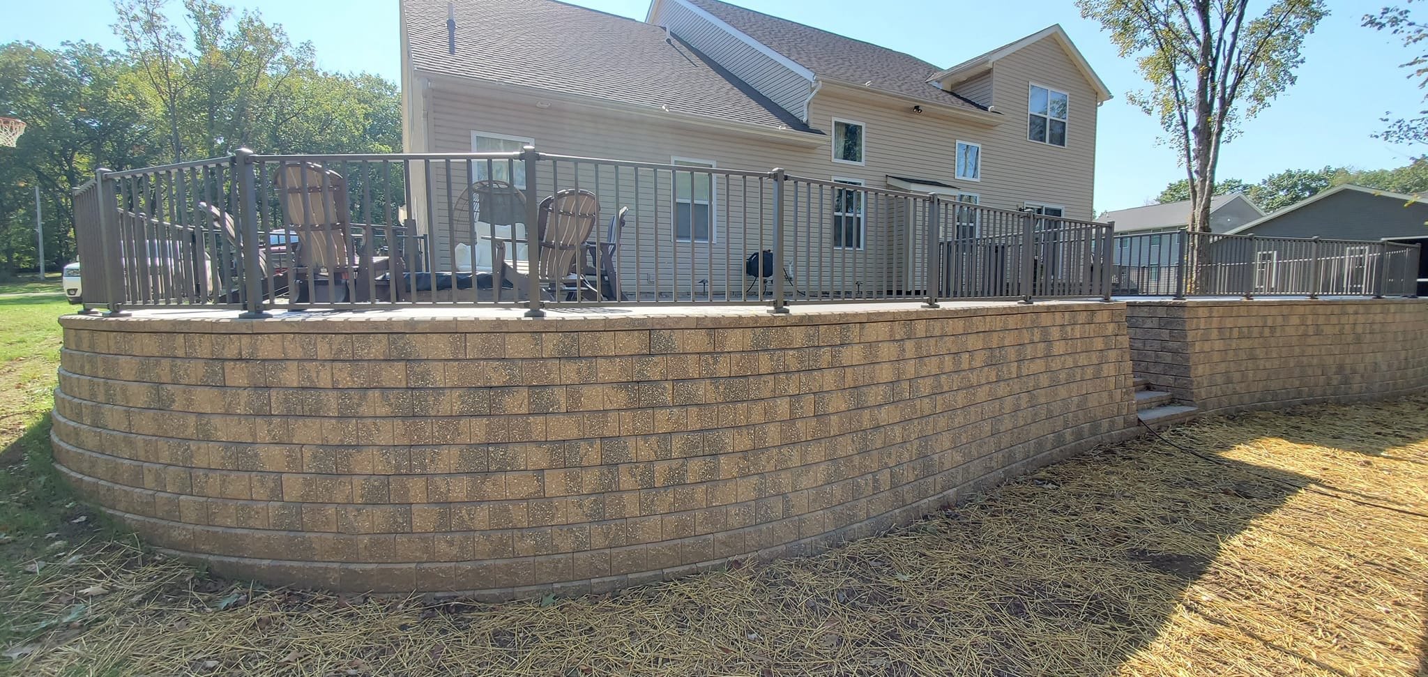 View of a backyard deck with metal railing and outdoor furniture, elevated on a brick retaining wall, with a beige house in the background.