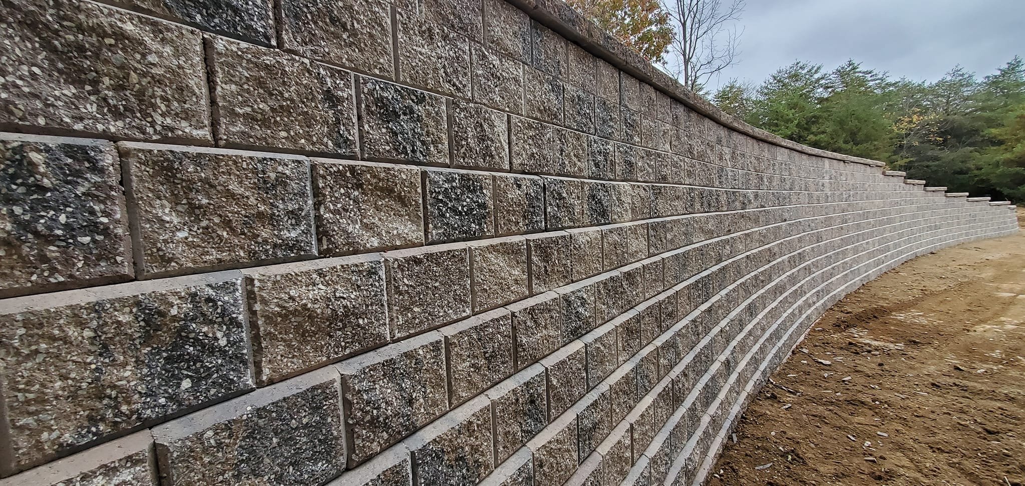 A long, curved brick retaining wall under construction, with trees in the background and dirt in the foreground.