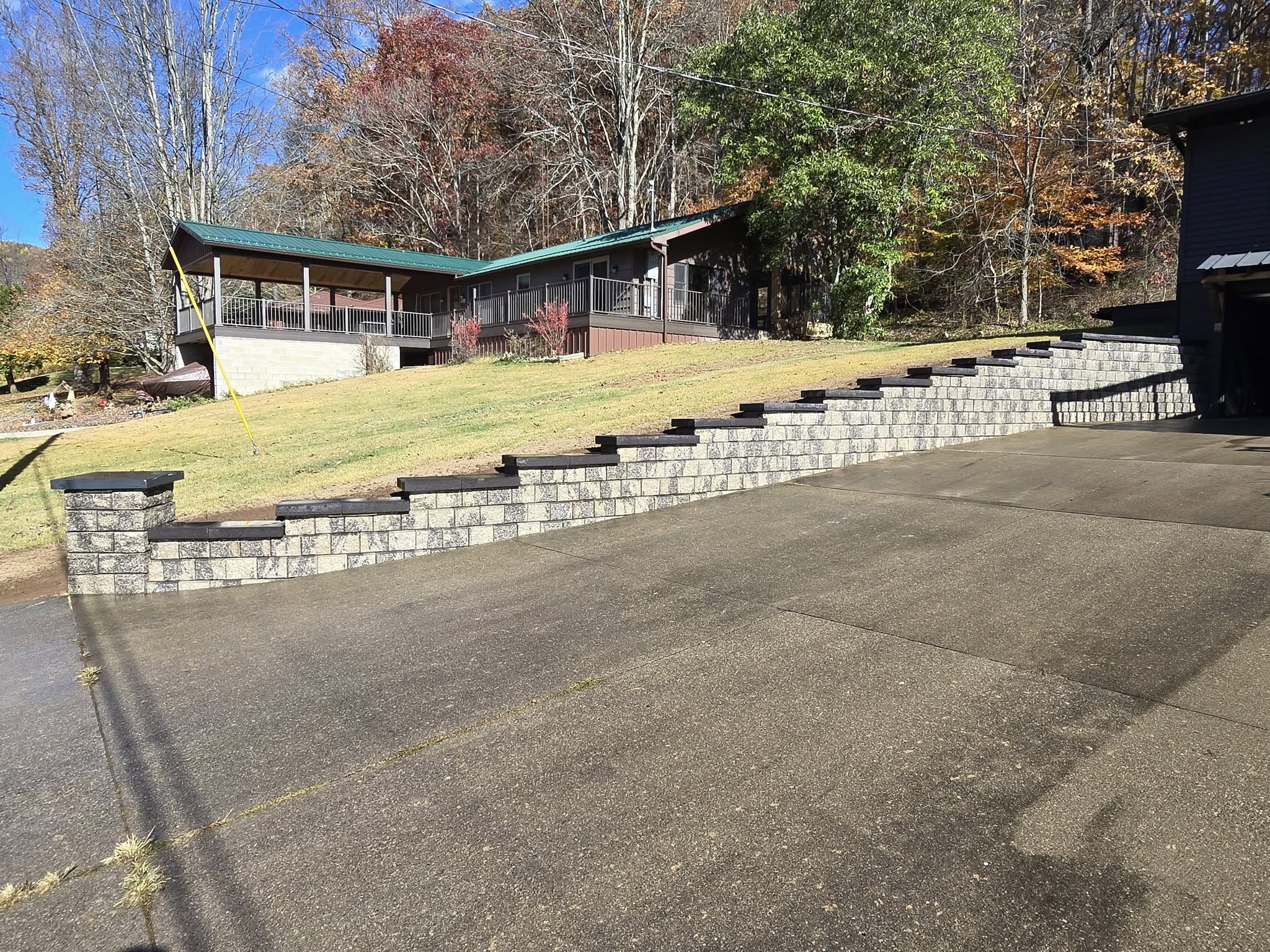 A stepped retaining wall made of gray bricks with dark caps, leading up to a grassy yard. In the background, there is a house with a covered porch and a green roof, situated on a hillside with trees showing autumn foliage.