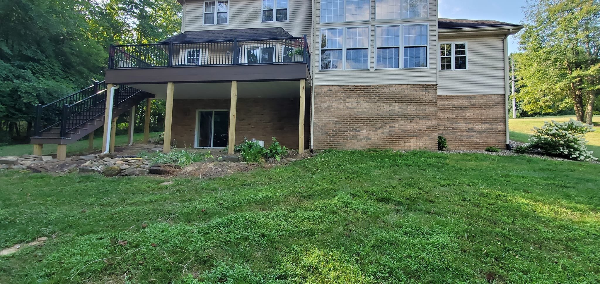 Backyard view of a multi-story house with a spacious elevated wooden deck. The house has brick and vinyl siding, and there is a garden bed with plants underneath the deck. A staircase on the left leads to the deck, and the yard is grassy with trees i