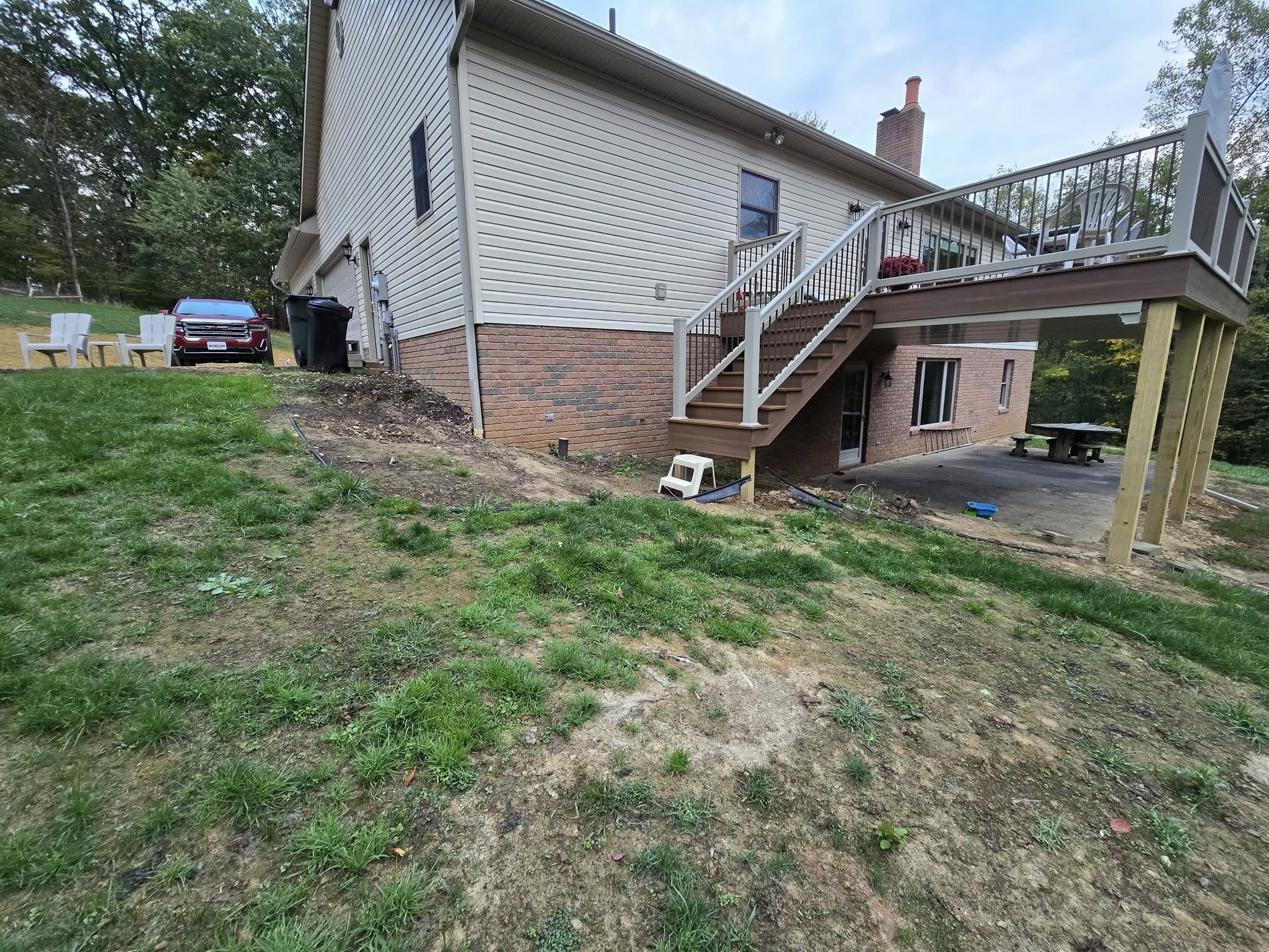 Backyard view of a house with a raised deck and stairs leading down to a partially landscaped yard. There are a few outdoor chairs on the deck, a white plastic chair at the bottom of the stairs, and some gardening or construction tools and materials 