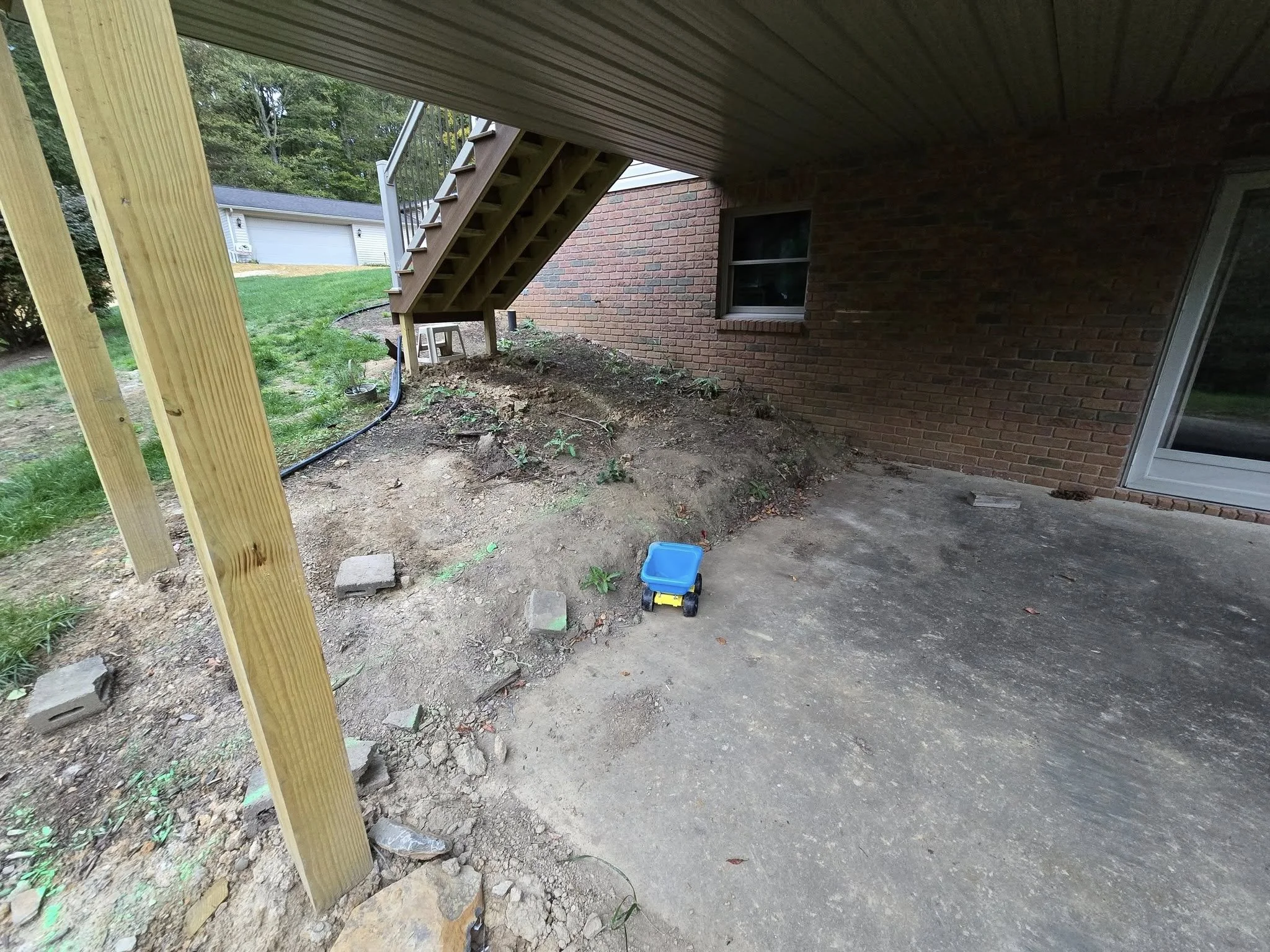 Backyard patio area with dirt and concrete ground, wooden staircase leading to upper level, brick house wall with a window and door, small toy truck on ground, green grass and trees in background.