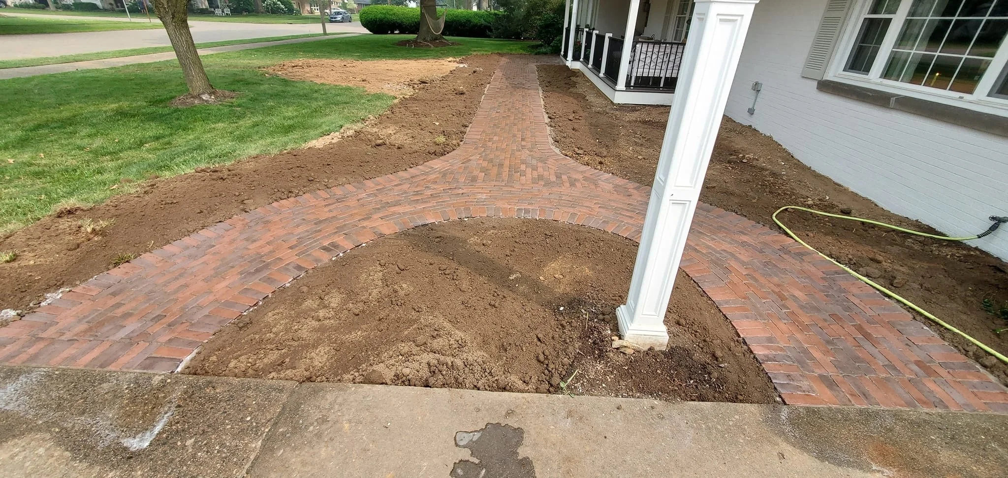 Newly constructed brick pathway curved around a porch area in a residential yard, with soil and grass areas surrounding it.