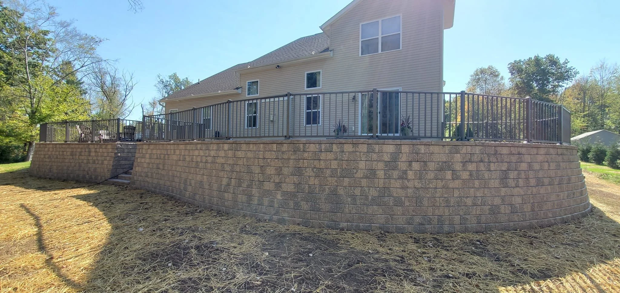 A large elevated backyard patio with a brick retaining wall and metal railing, attached to a beige house with multiple glass doors and windows, surrounded by trees and a clear blue sky.