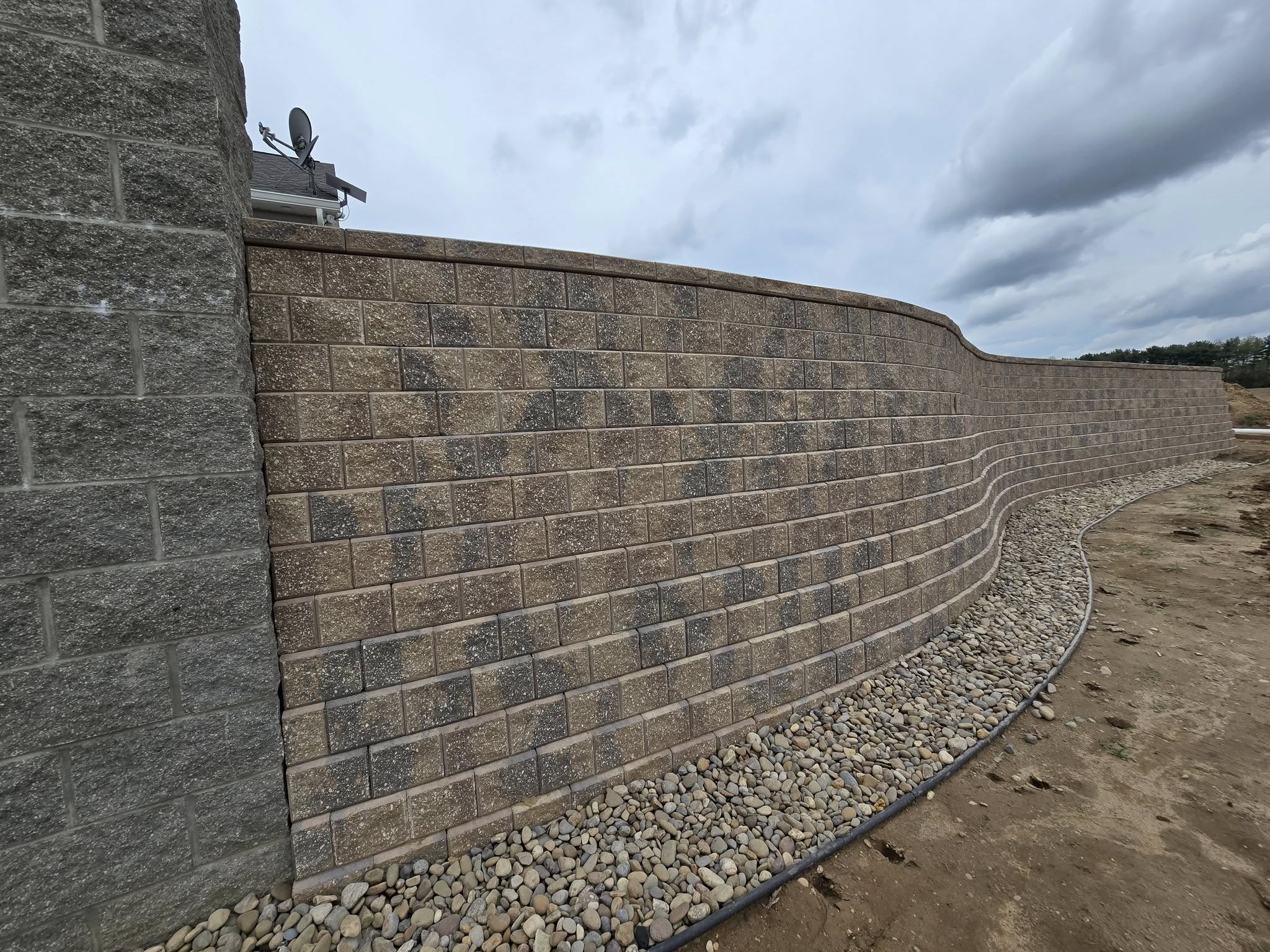 A curved brick retaining wall with a layer of small rounded rocks at its base, and a cloudy sky overhead, in an outdoor construction site.