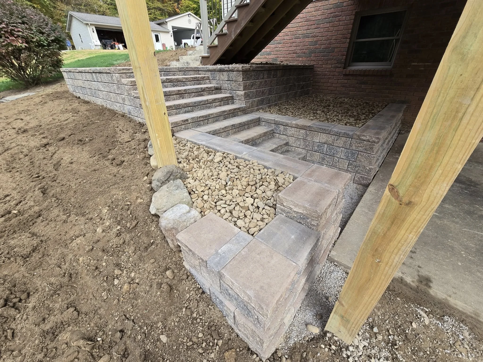 Newly constructed brick retaining wall and stairs outside a house. Loose gravel and rocks are on the ground in front of the wall. Wooden posts support a raised deck or porch above.