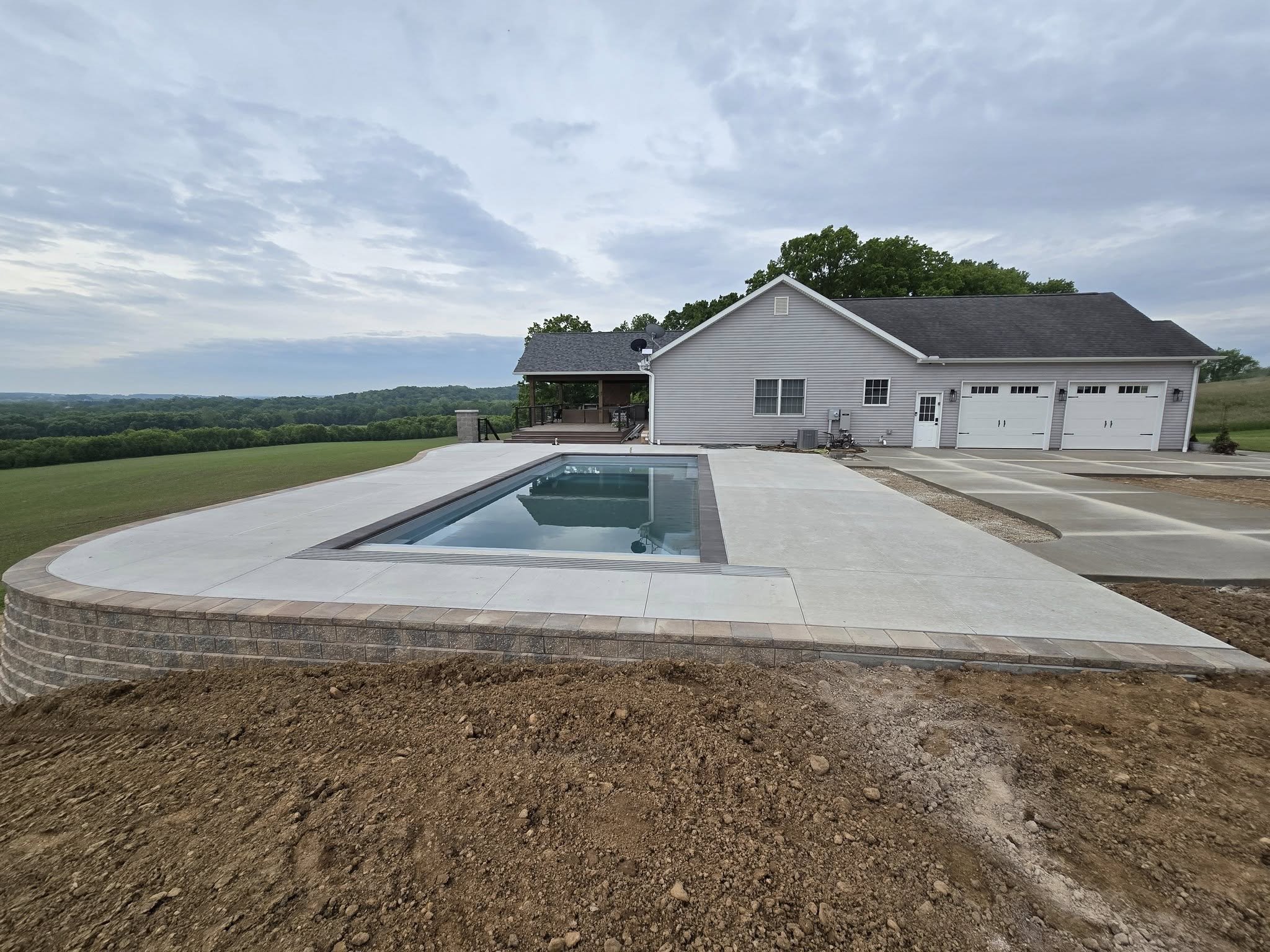 Backyard with in-ground swimming pool, patio, house, and open countryside with cloudy sky.