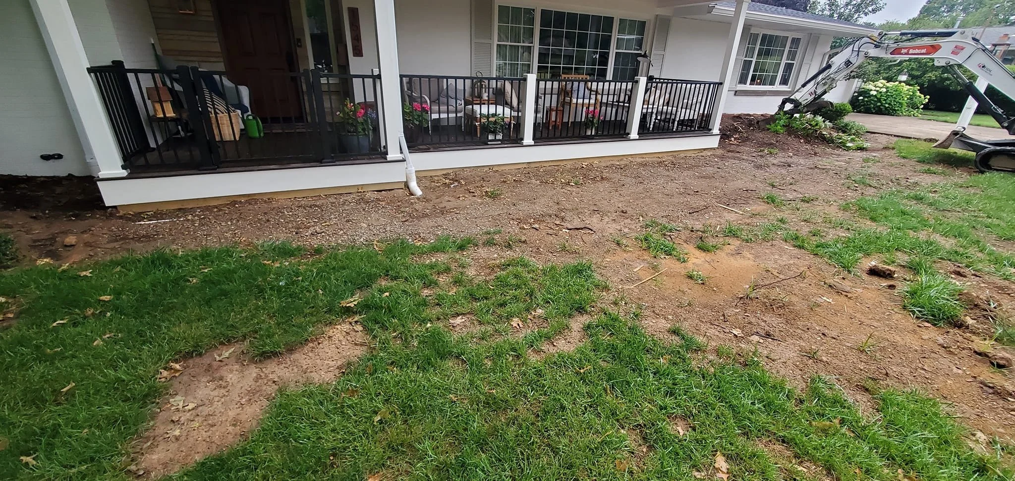 A house with a screened porch under construction, with patches of dirt and grass in the yard and a small construction excavator on the right side.