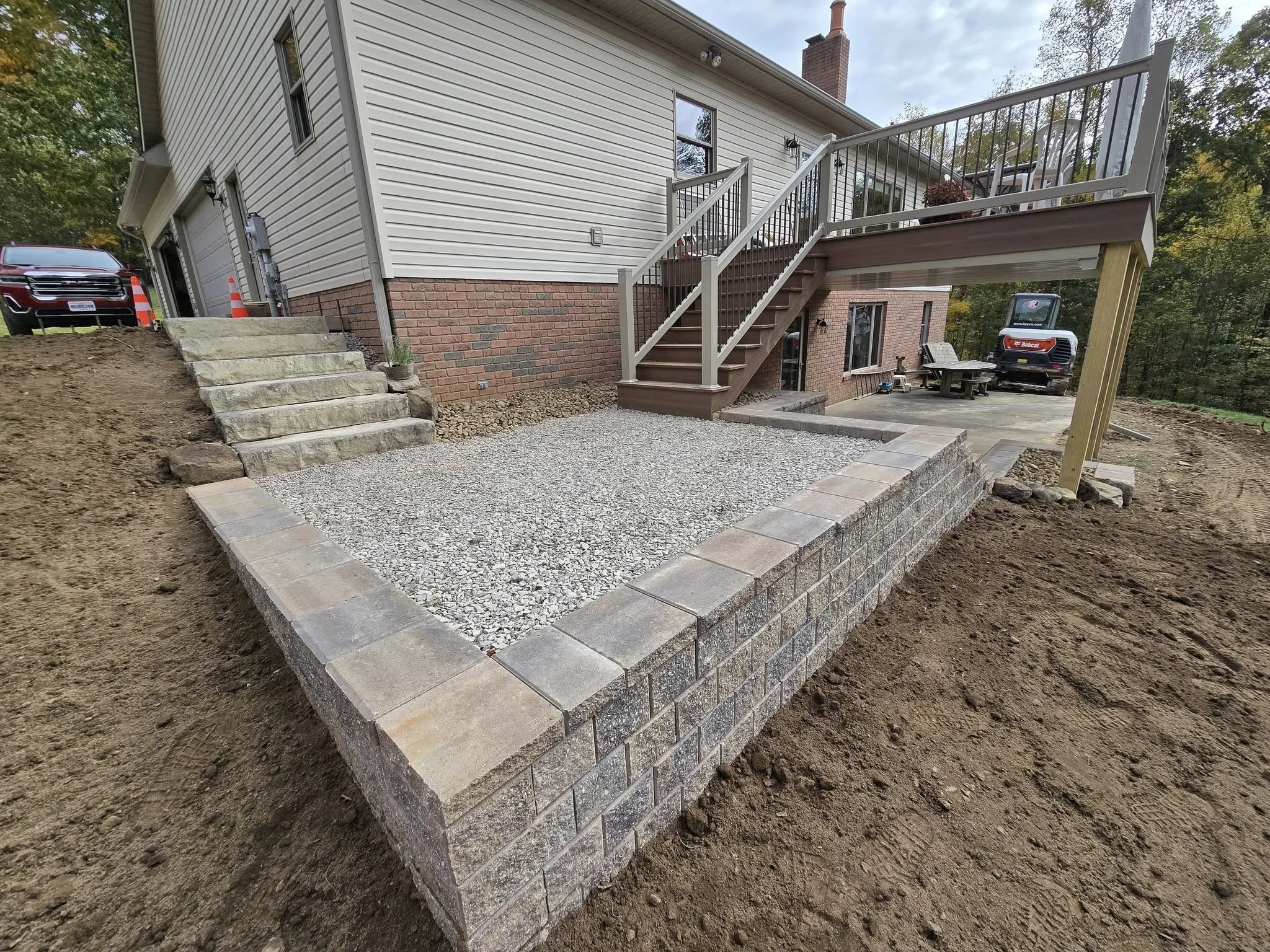 Backyard construction with newly built raised deck, gravel patio, stone steps, and retaining wall, with construction vehicles and tools in the background.