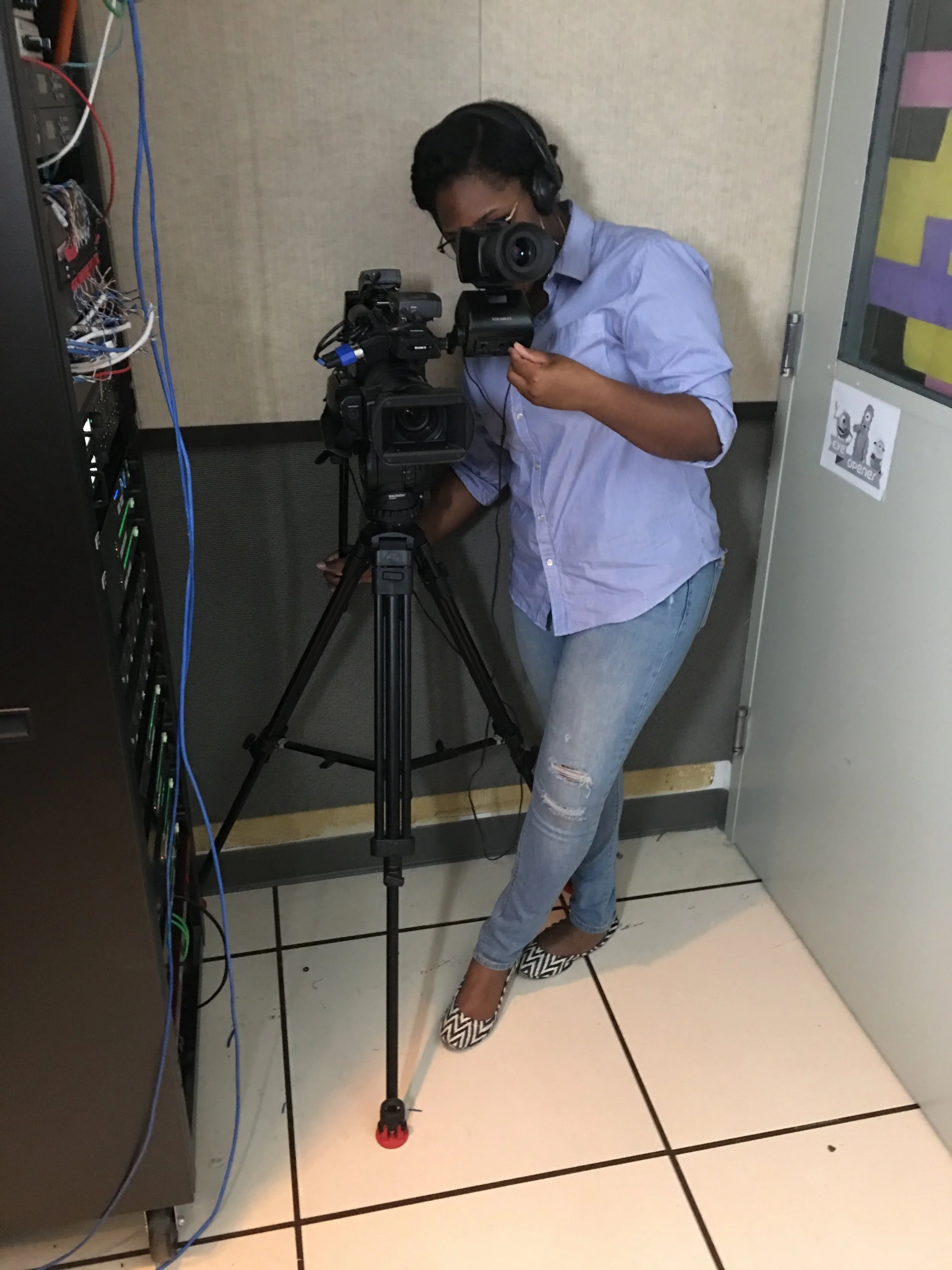 A person filming with a professional video camera on a tripod in a small room, wearing headphones and jeans with ripped knees.