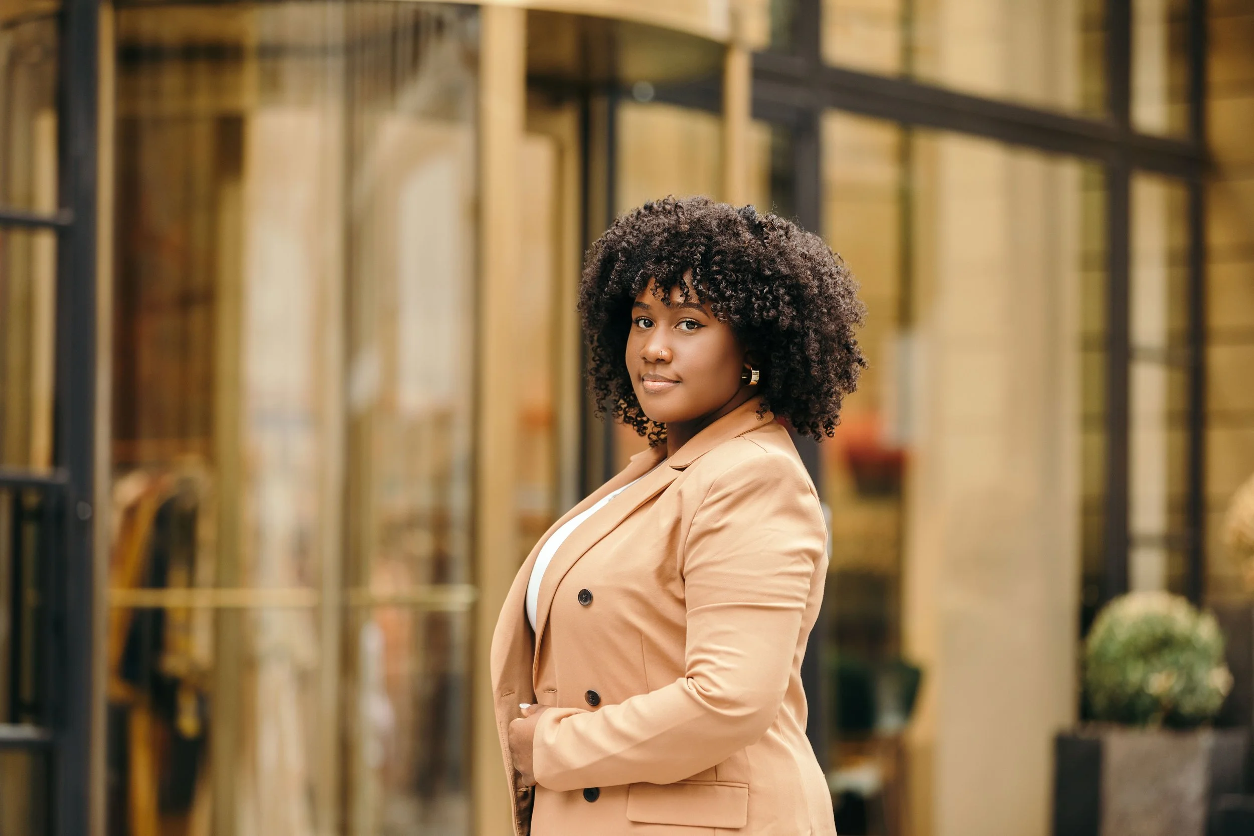 A woman with curly hair wearing a beige blazer standing outdoors near a glass building.