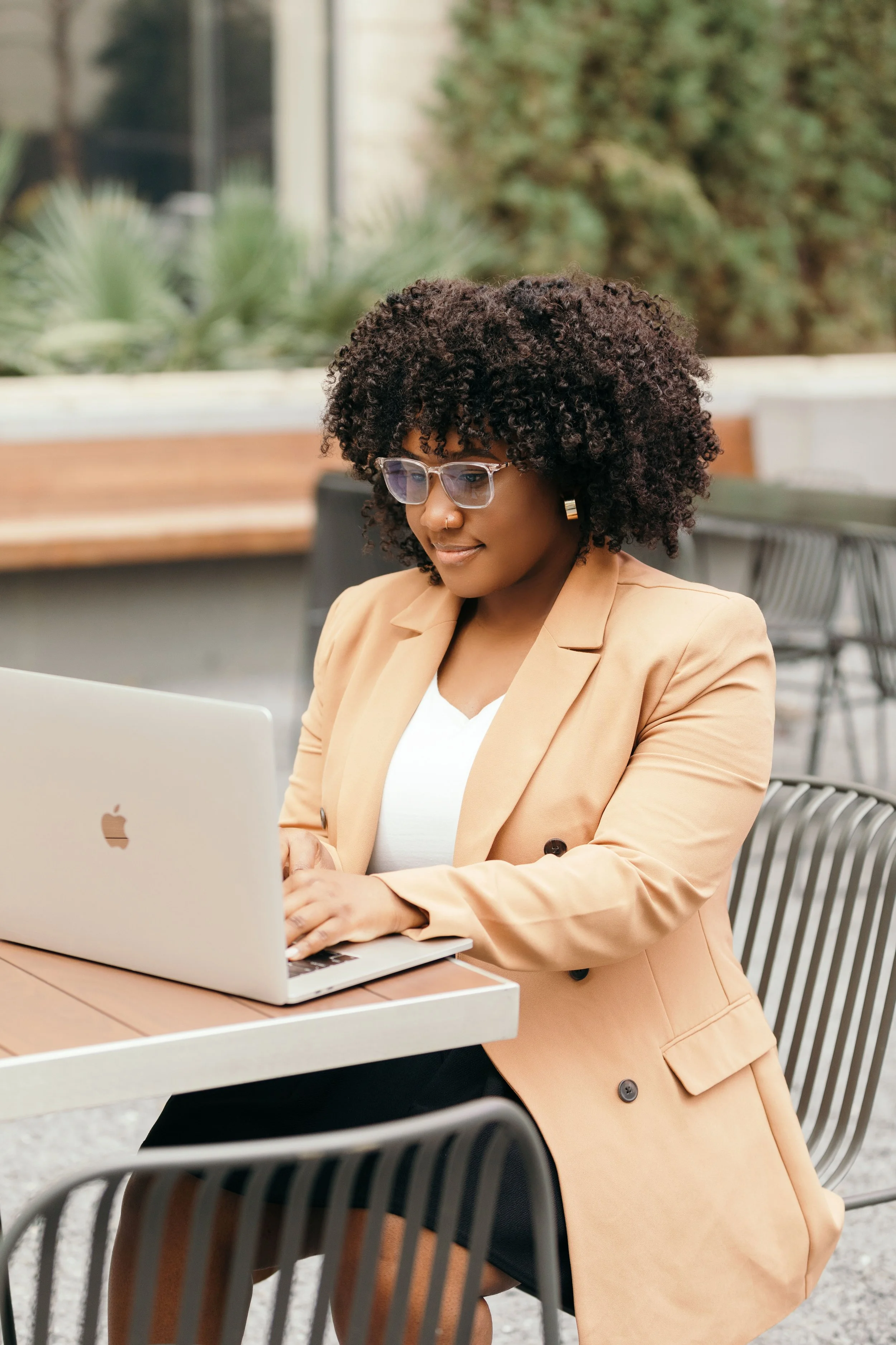 A woman with curly hair, glasses, and a beige blazer working on a silver MacBook at an outdoor table.