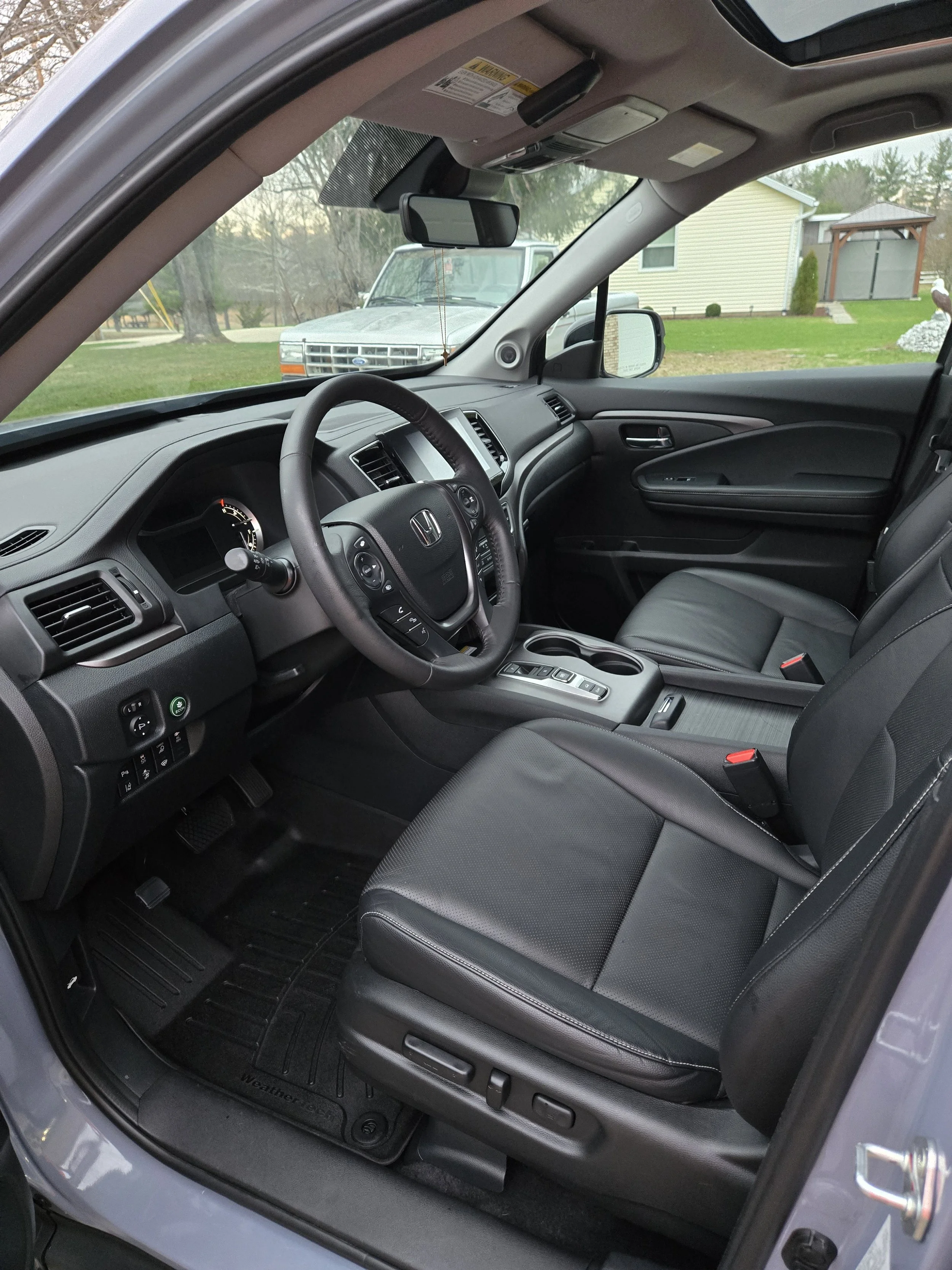 Interior view of a Honda vehicle with black seats and dashboard, showing the steering wheel, gear shift, and front window, with a house and trees visible outside.