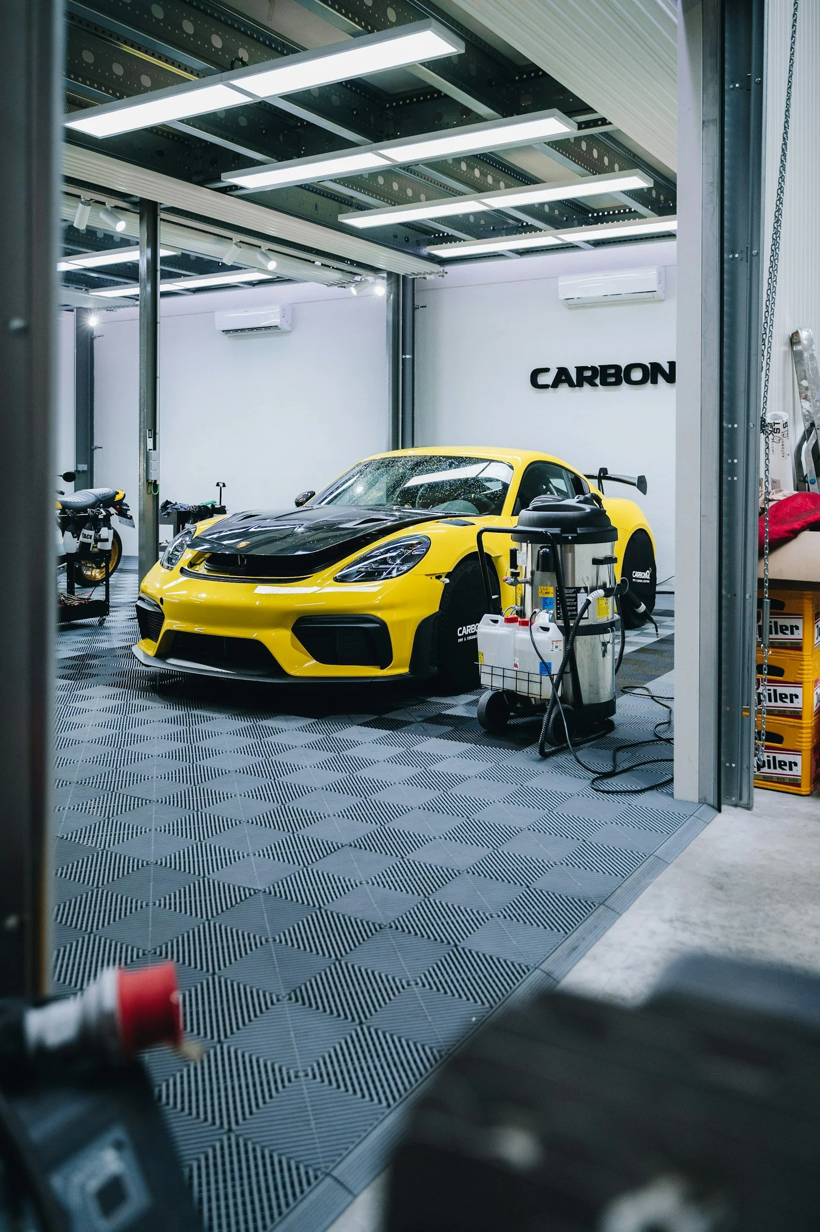 Yellow sports car being cleaned inside a workshop with the word 'CARBON' on the wall.