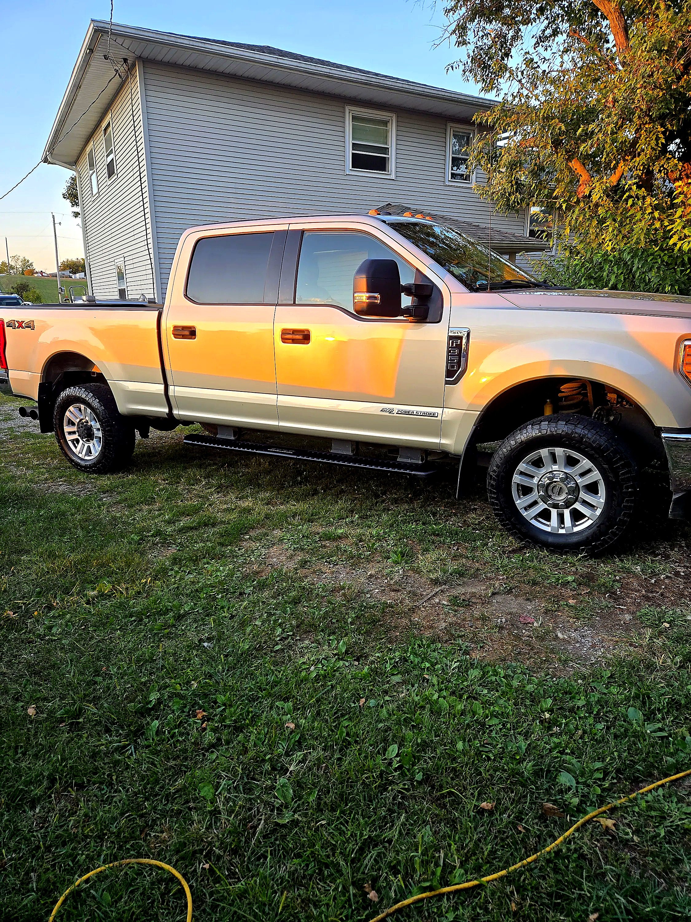 A beige and orange Ford F-250 pickup truck parked on a grassy area near a light blue house with multiple windows and trees, during sunset.