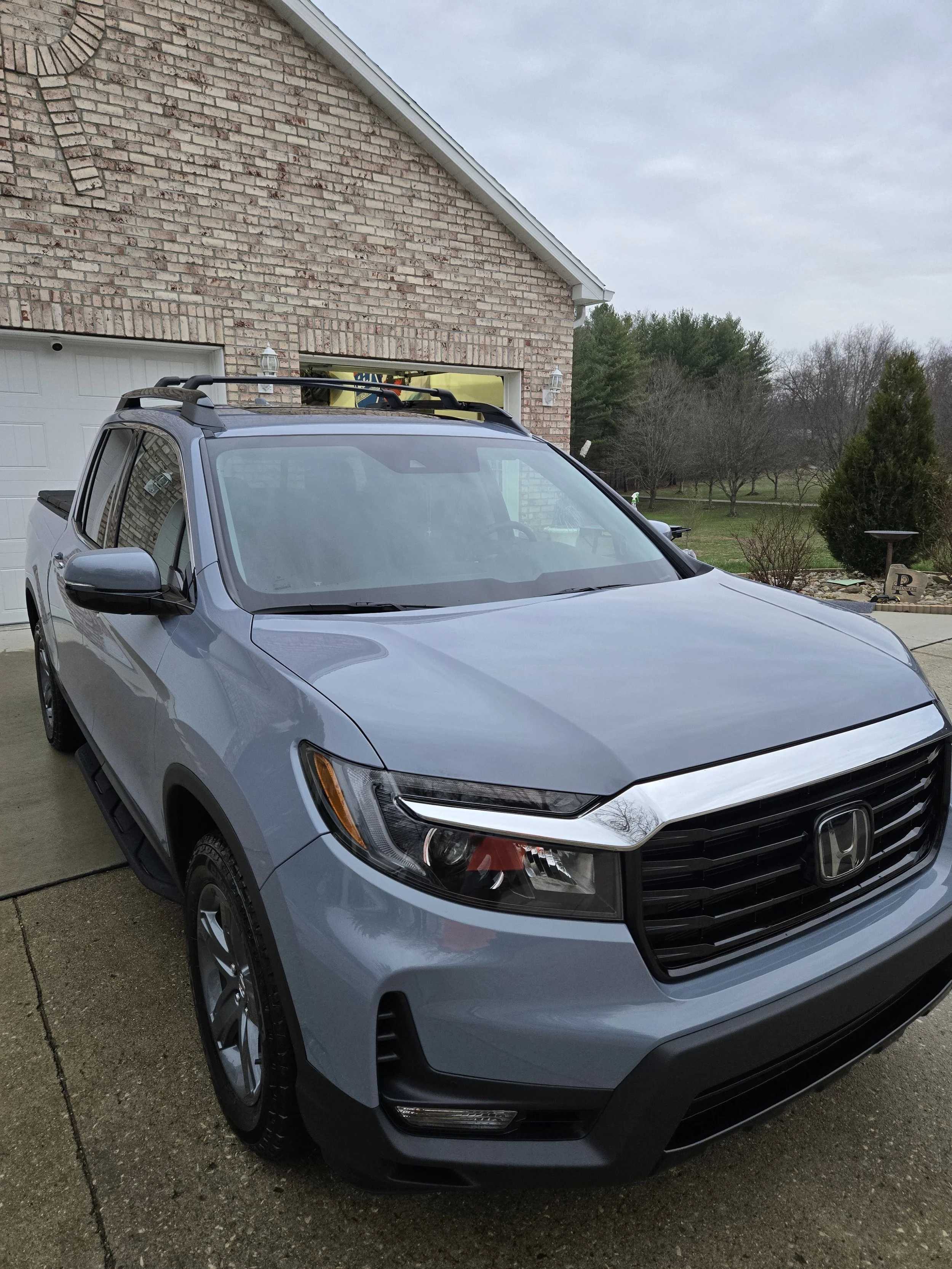 A silver Honda SUV parked in front of a brick house with a garage door, on a concrete driveway. The house has a sloped roof and outdoor lights on the wall.