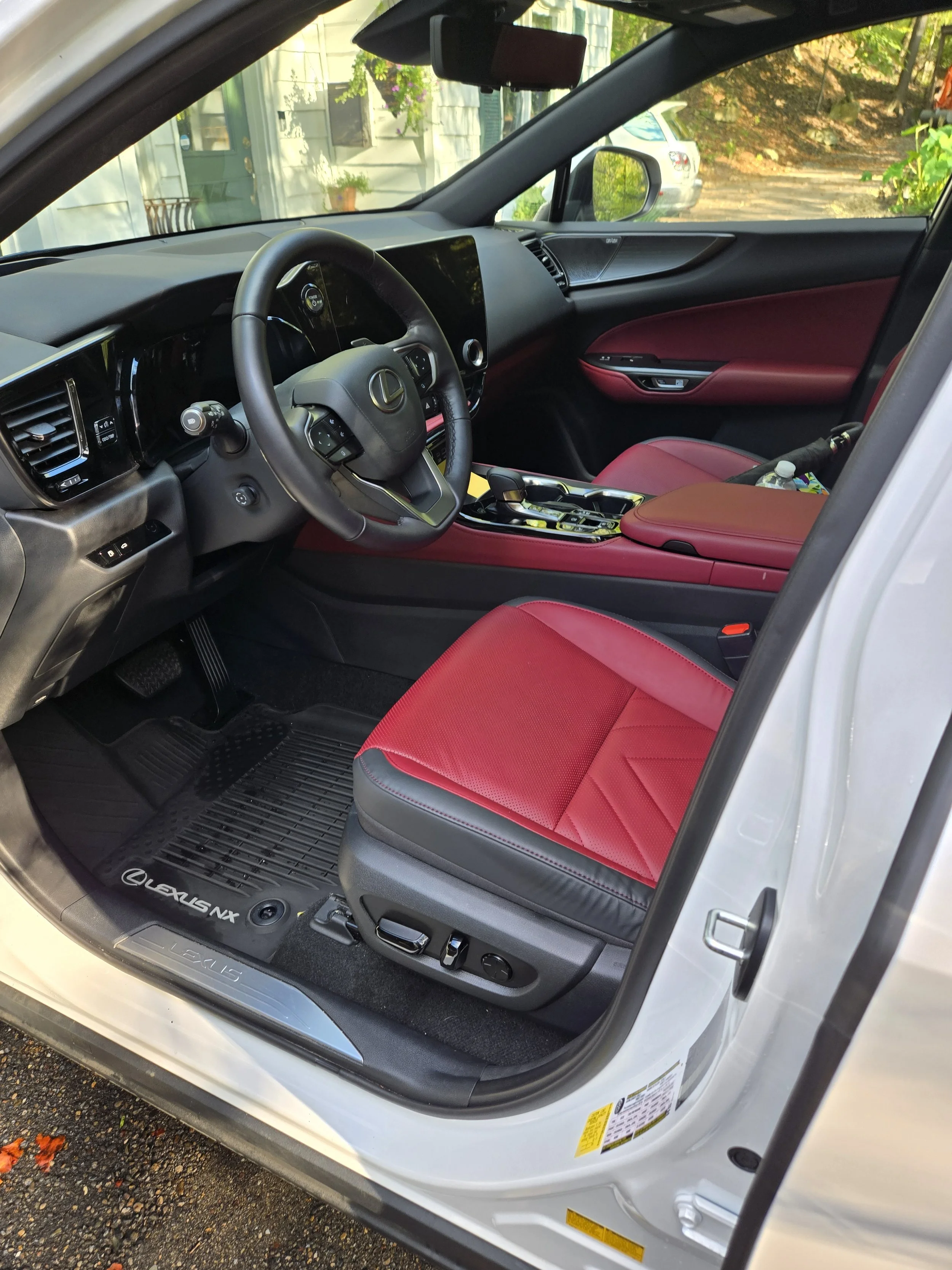 The interior of a white Lexus vehicle with red leather seats, black dashboard, and steering wheel, parked outside near greenery, with passenger side door open.