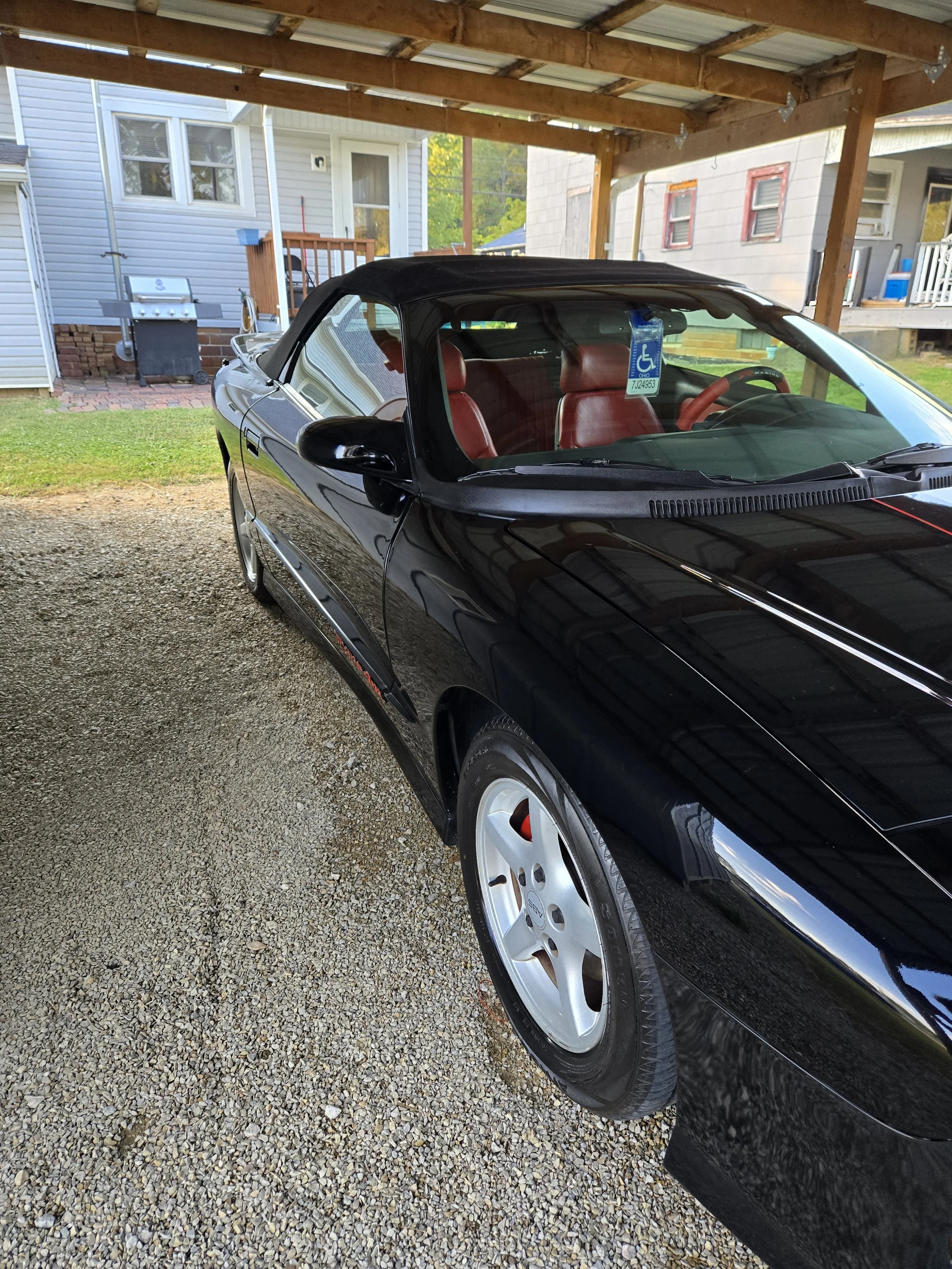 A black convertible car parked under a carport on gravel, with a red interior and a handicap parking permit hanging from the mirror.