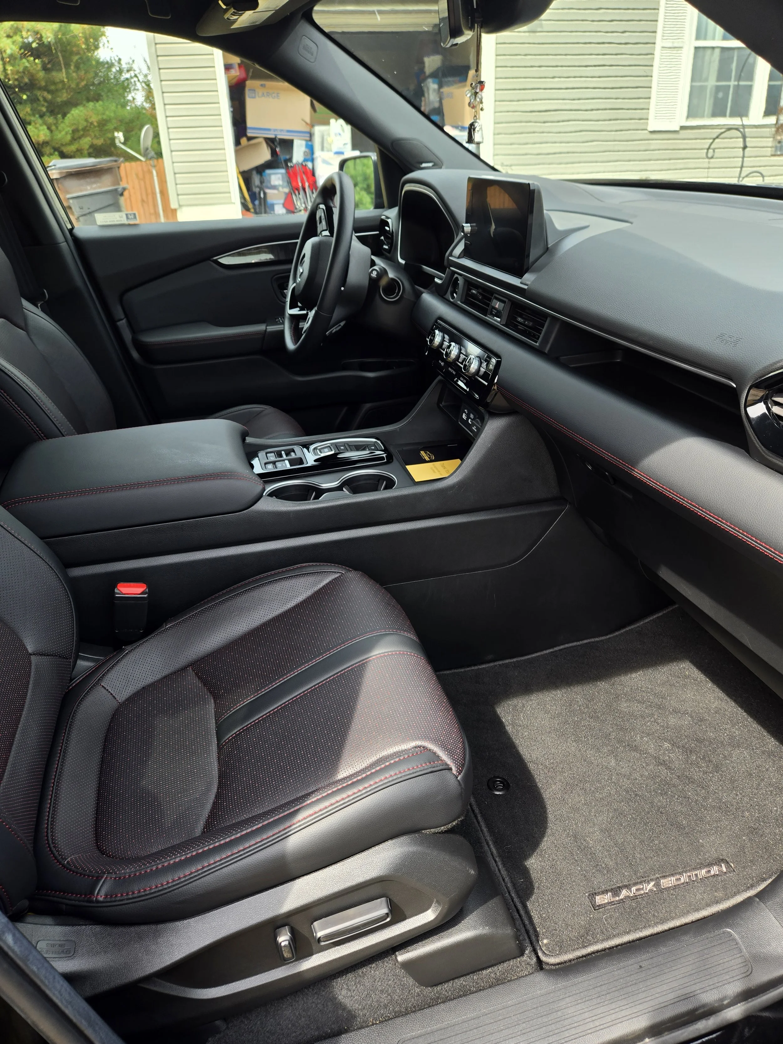 Interior of a modern car featuring black leather seats with red stitching, a dashboard with a digital display, and a center console with controls. The driver seat has electronic adjustment buttons, and there is a branded black floor mat with "BLACK E