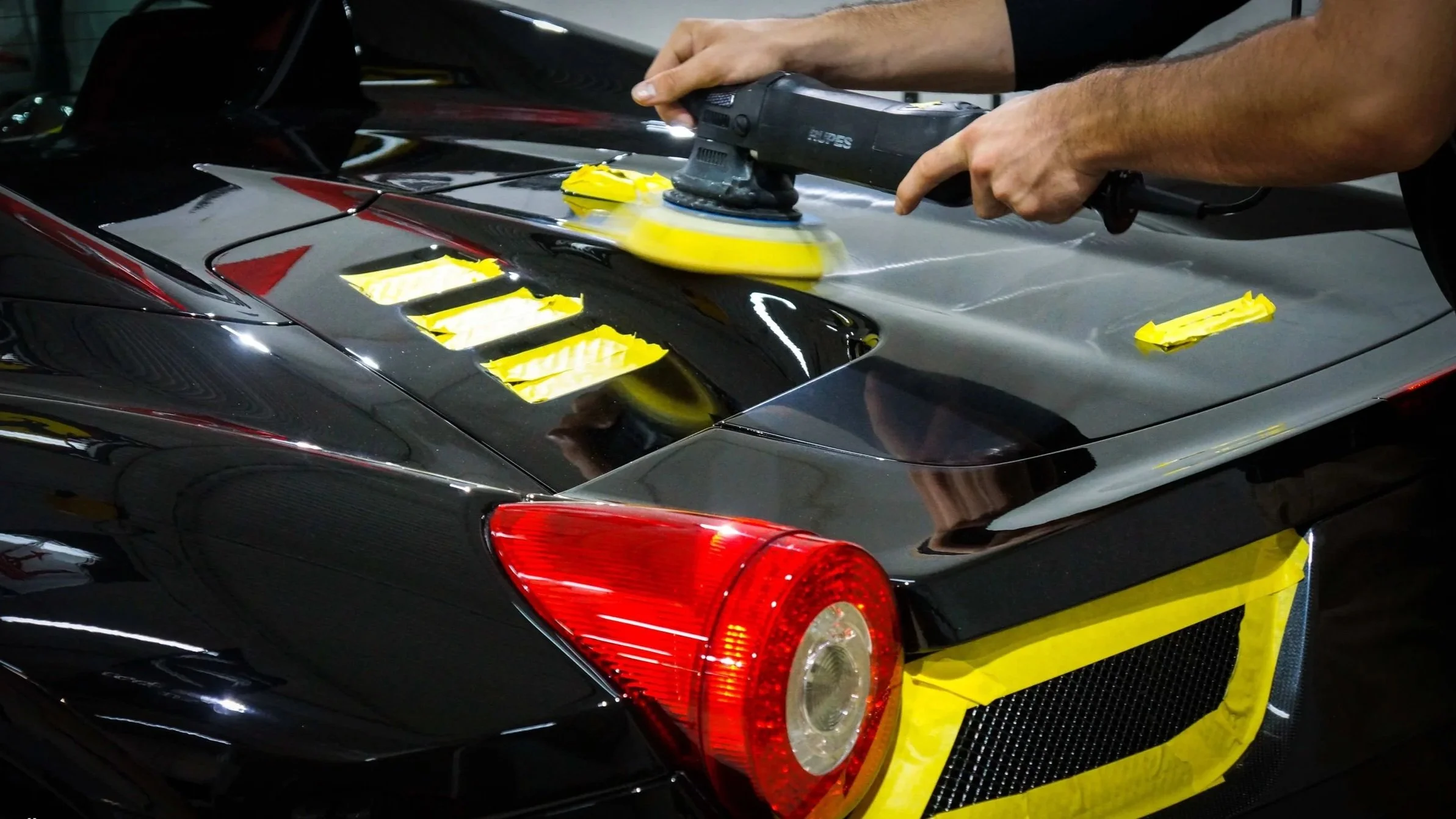 Person polishing the black and red sports car with a yellow polishing machine, yellow tape partially covering the rear of the car.