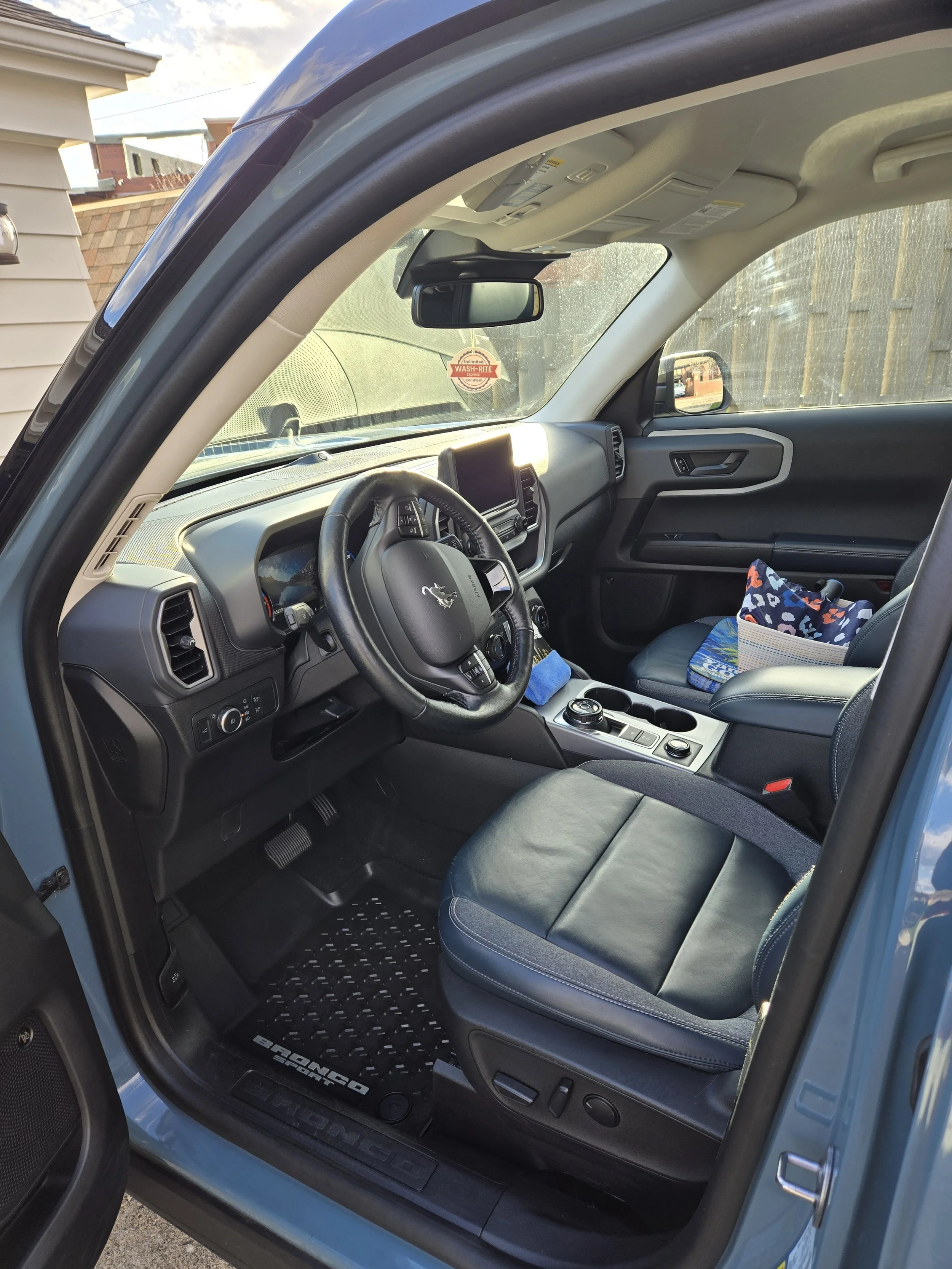 Interior of a vehicle showing the driver's seat, steering wheel, dashboard, and front passenger seat with a colorful blanket and box on it.