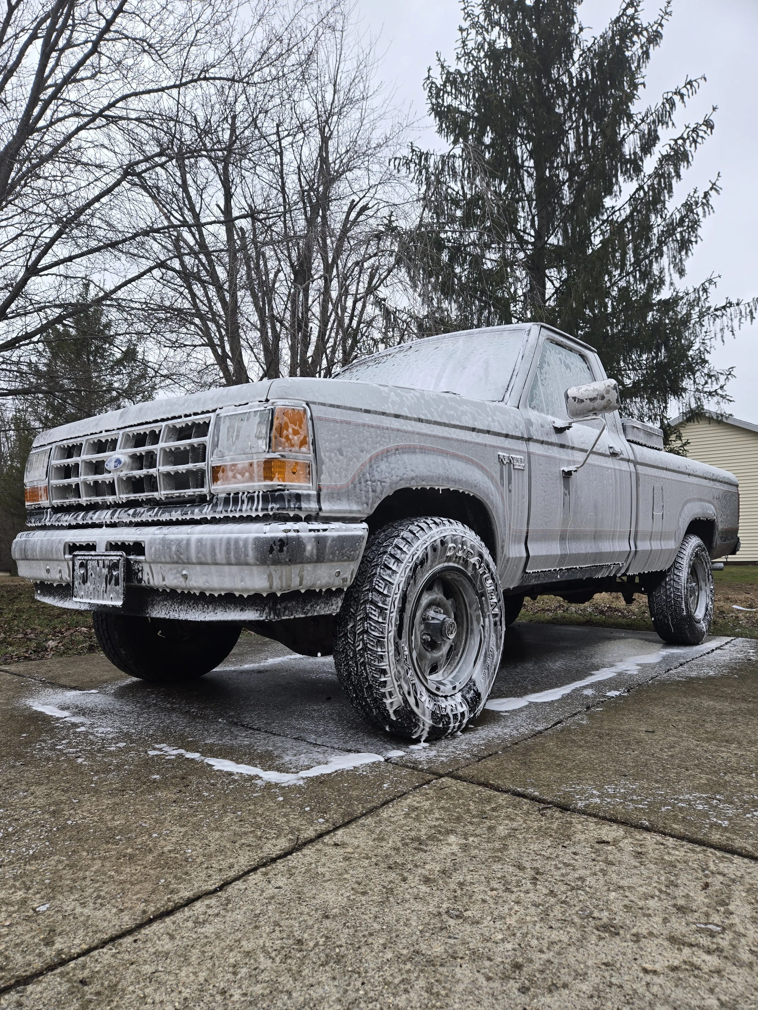 A silver pickup truck covered in soap and foam, parked on a driveway during a cloudy day, with leafless trees and a house in the background.