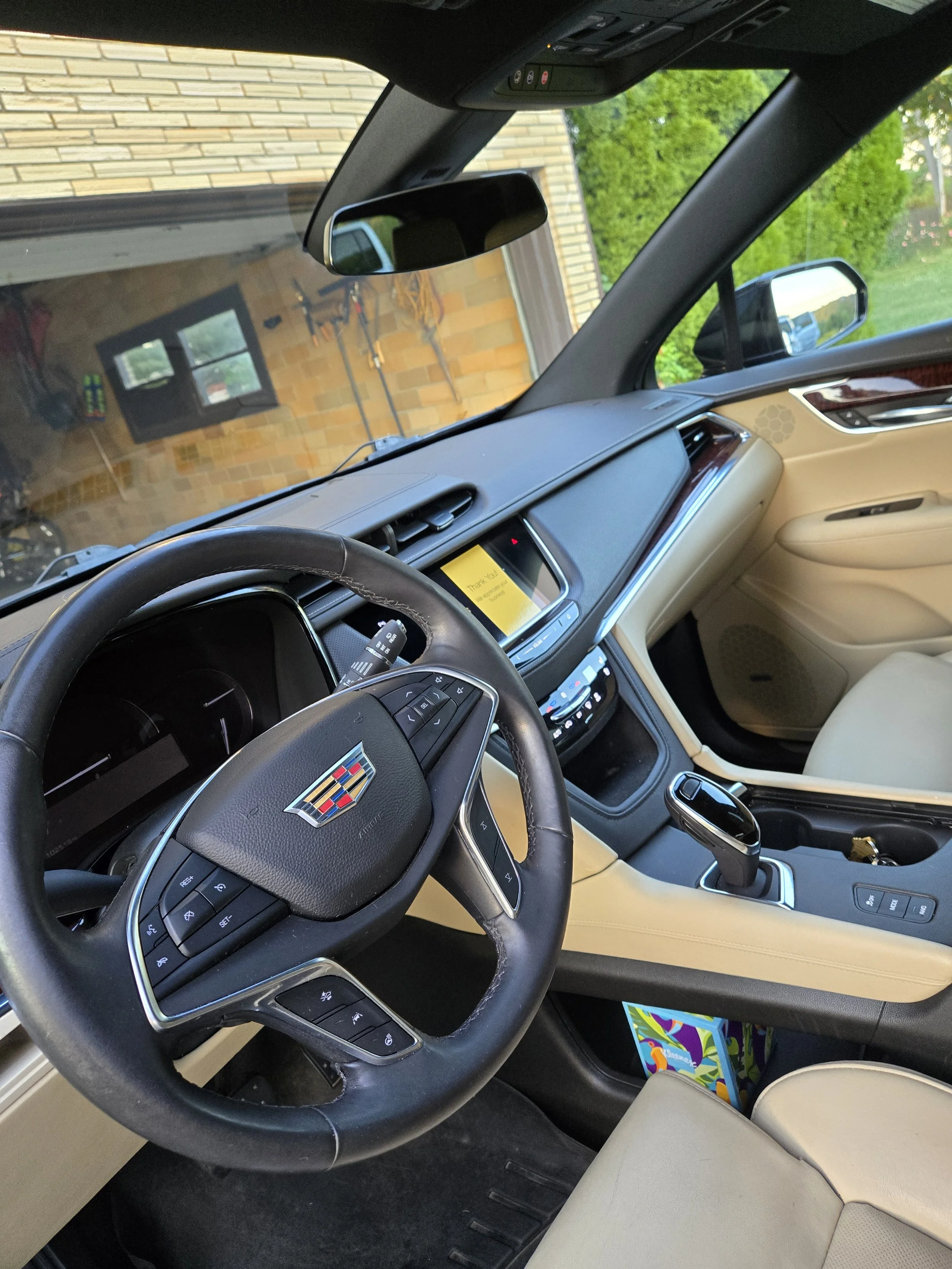 Interior of a Cadillac vehicle, showing the steering wheel, dashboard, gear shifter, and front seats, with a view of a garage and a house outside through the windshield.