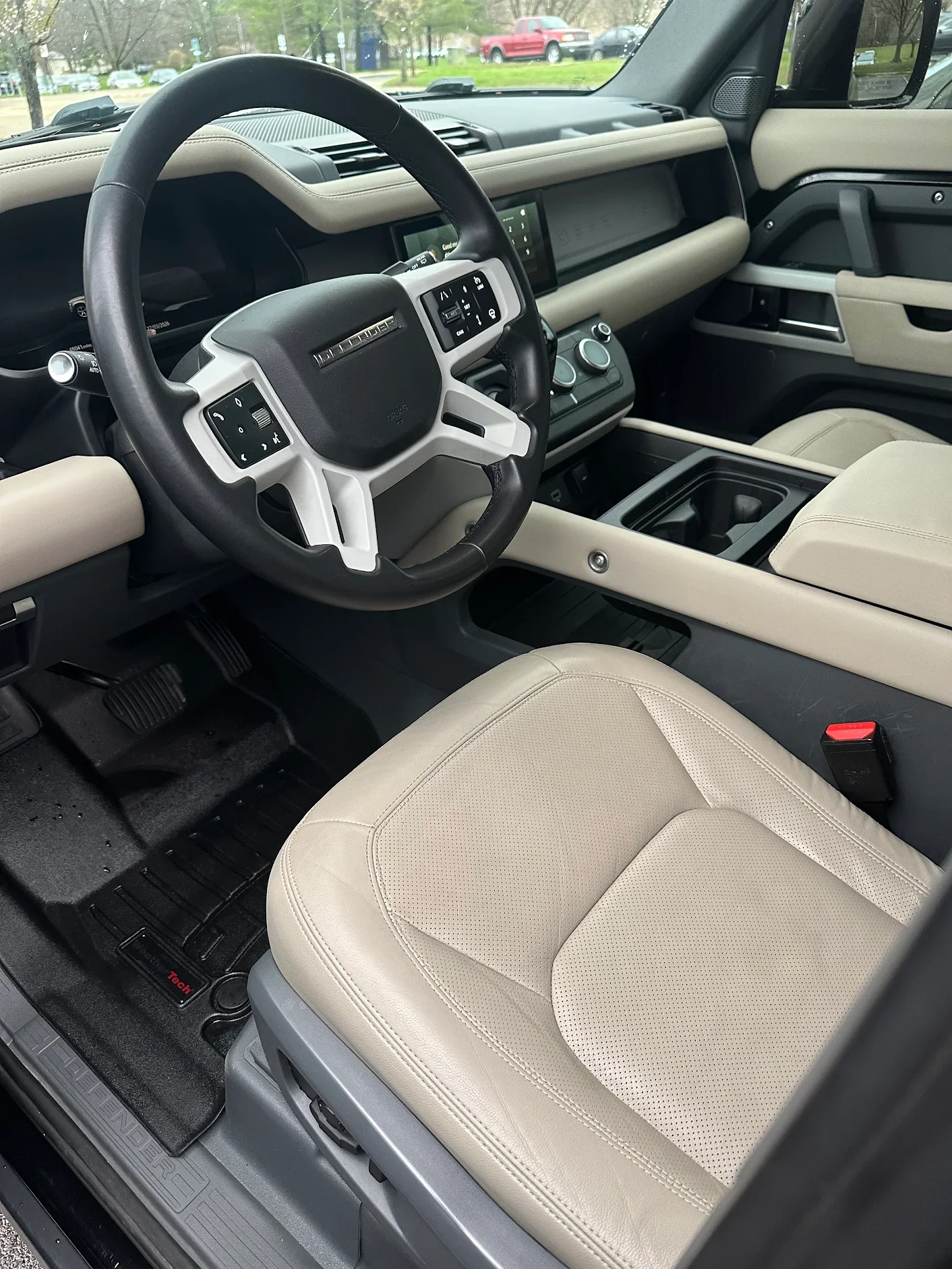 Interior view of a vehicle's front driver seat area, showing a beige seat, steering wheel, dashboard with controls, and center console.